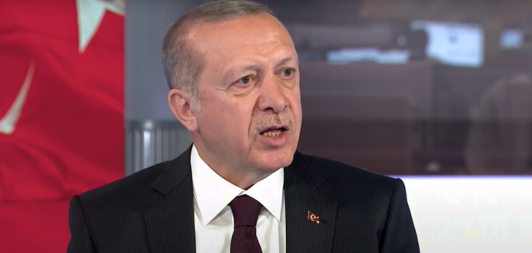 Erdogan with short gray hair, wearing a dark pinstripe suit, white shirt, and maroon tie, speaking with a Turkish flag in the background.