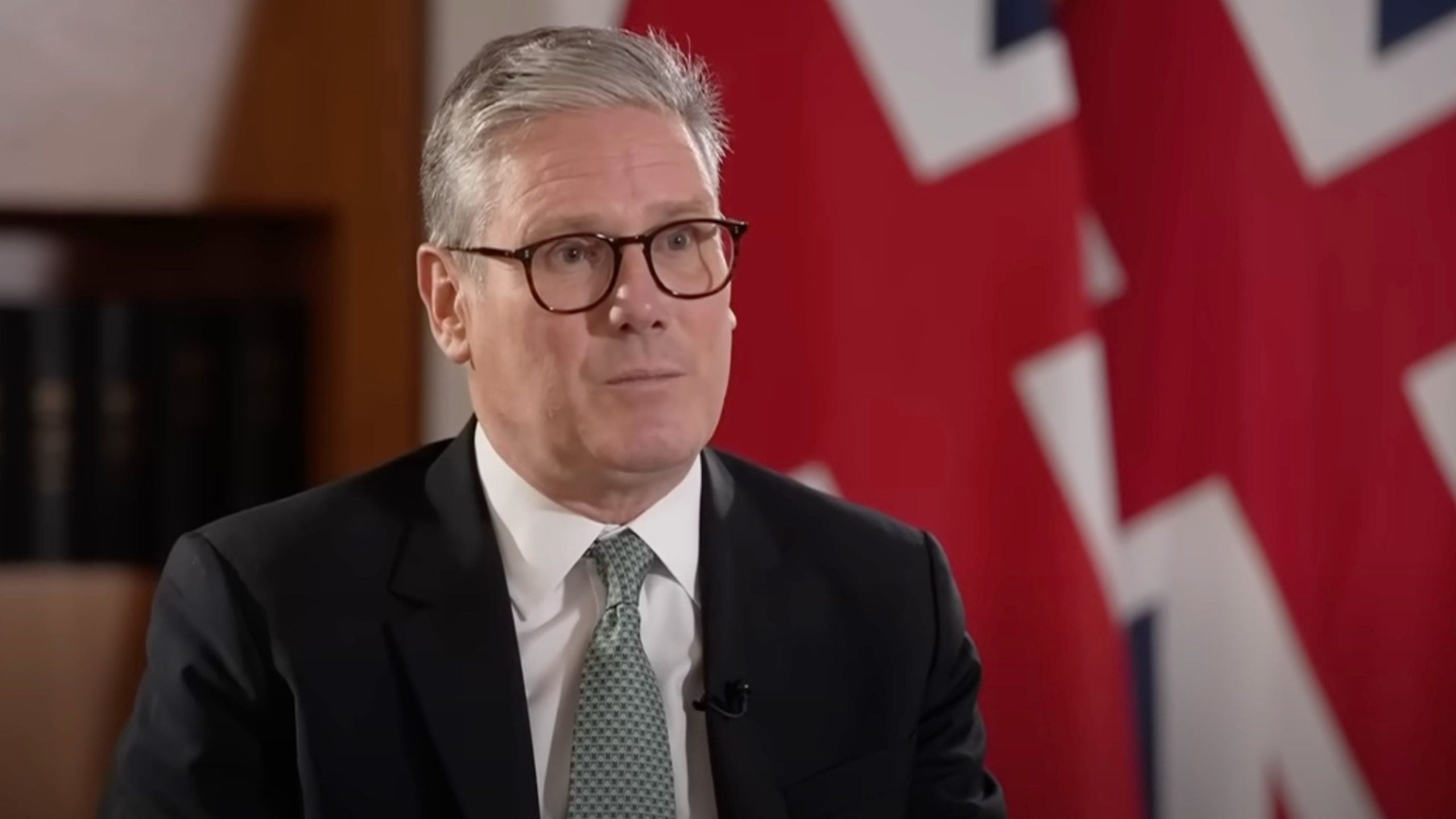Starmer in a suit and glasses speaking on camera, with a blurred British flag in the background.