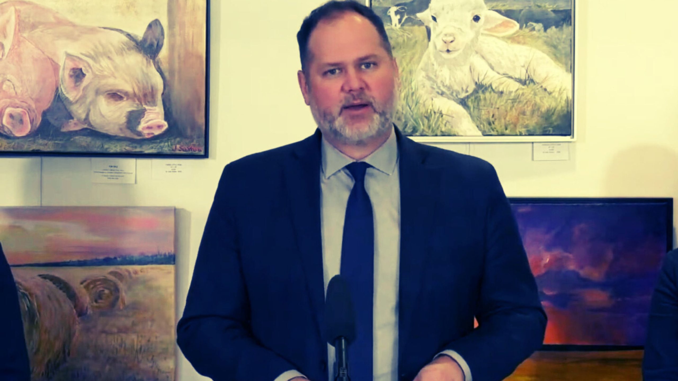 Matt Wiebe in a suit speaking at a podium, with paintings of animals and landscapes on the wall behind him.