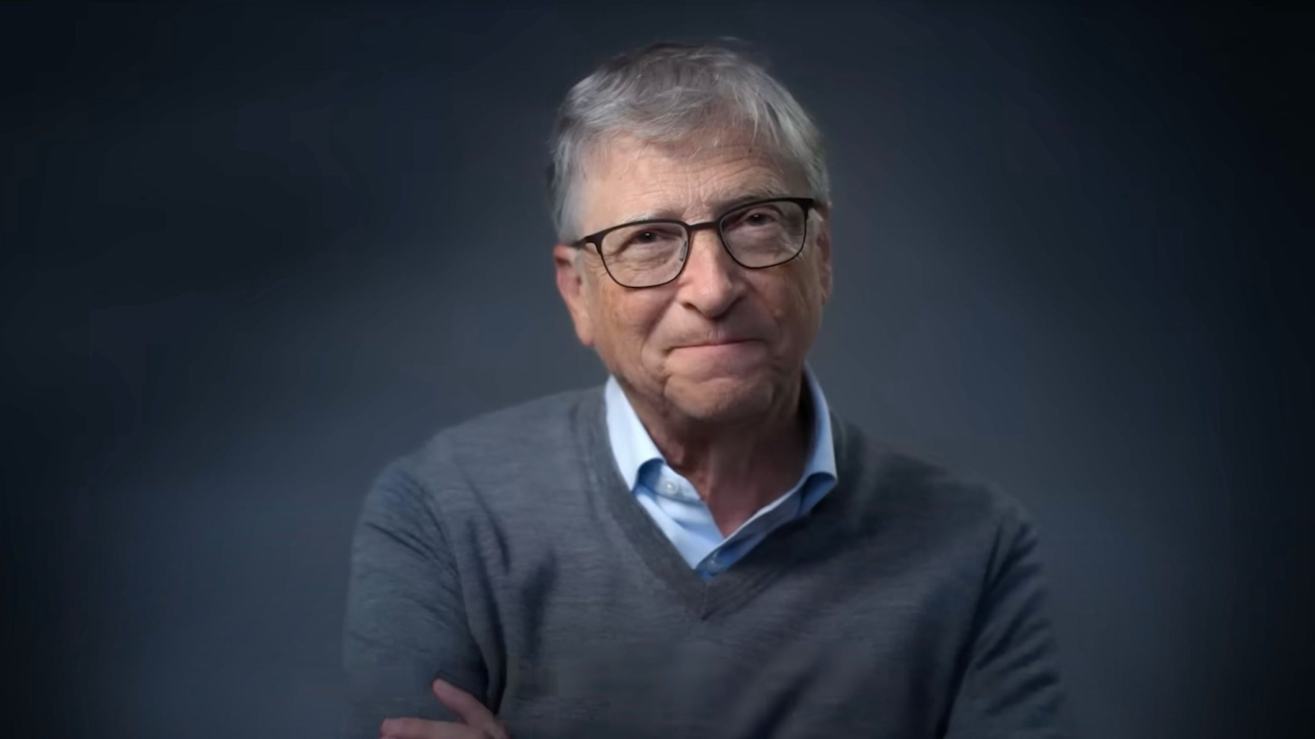 Bill Gates with short gray hair and glasses smiling, wearing a gray sweater over a light blue shirt against a dark background.