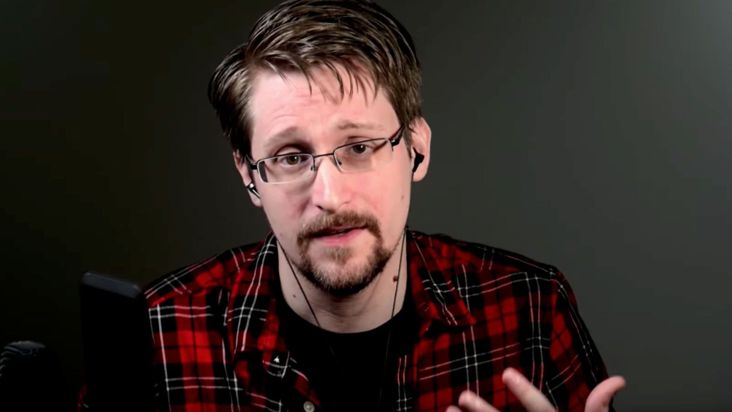 Edward Snowden with glasses wearing a red plaid shirt is speaking, with a microphone visible in front.