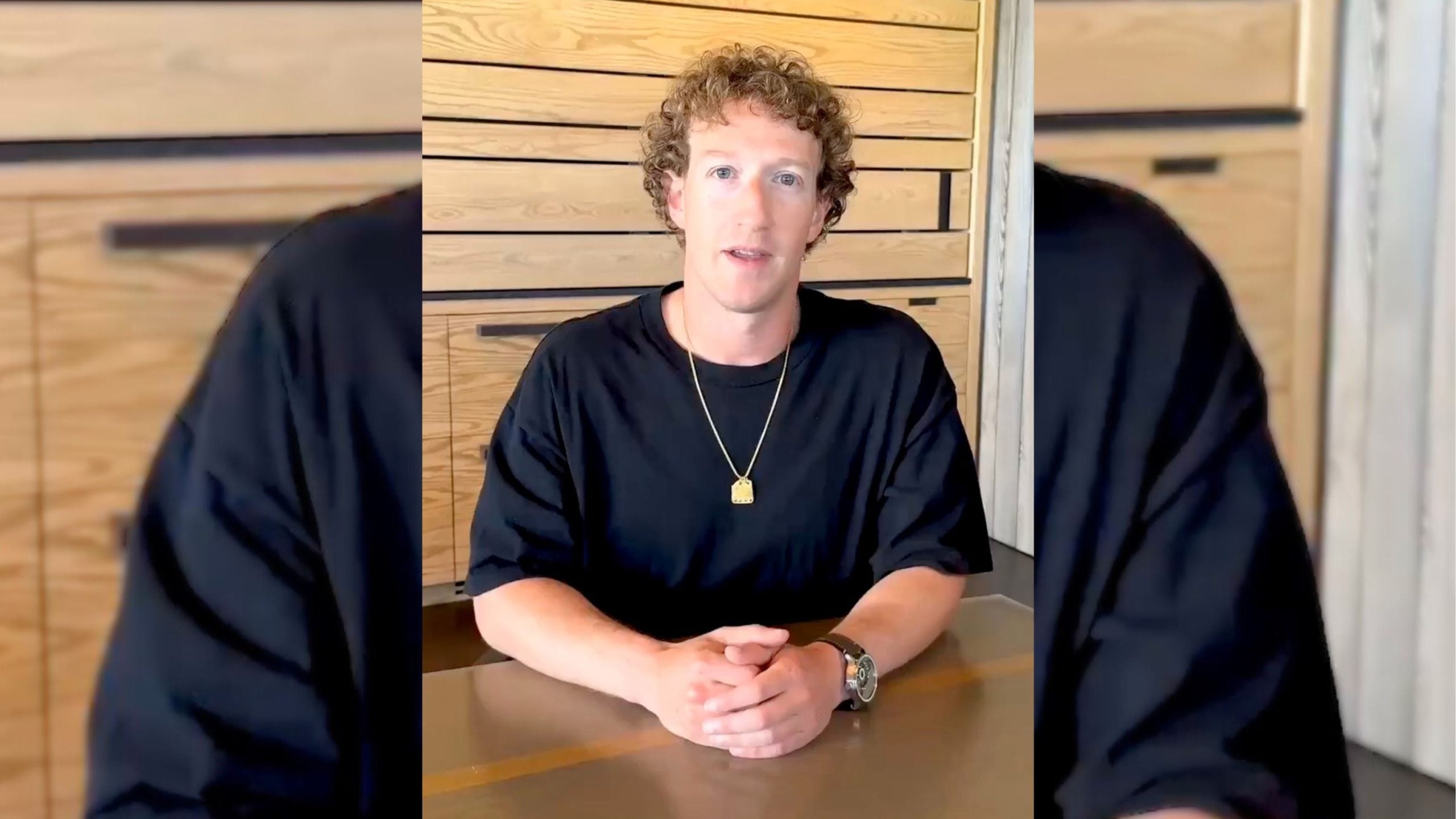 Zuckerberg with curly hair wearing a black shirt and a gold necklace sits at a table, hands folded.