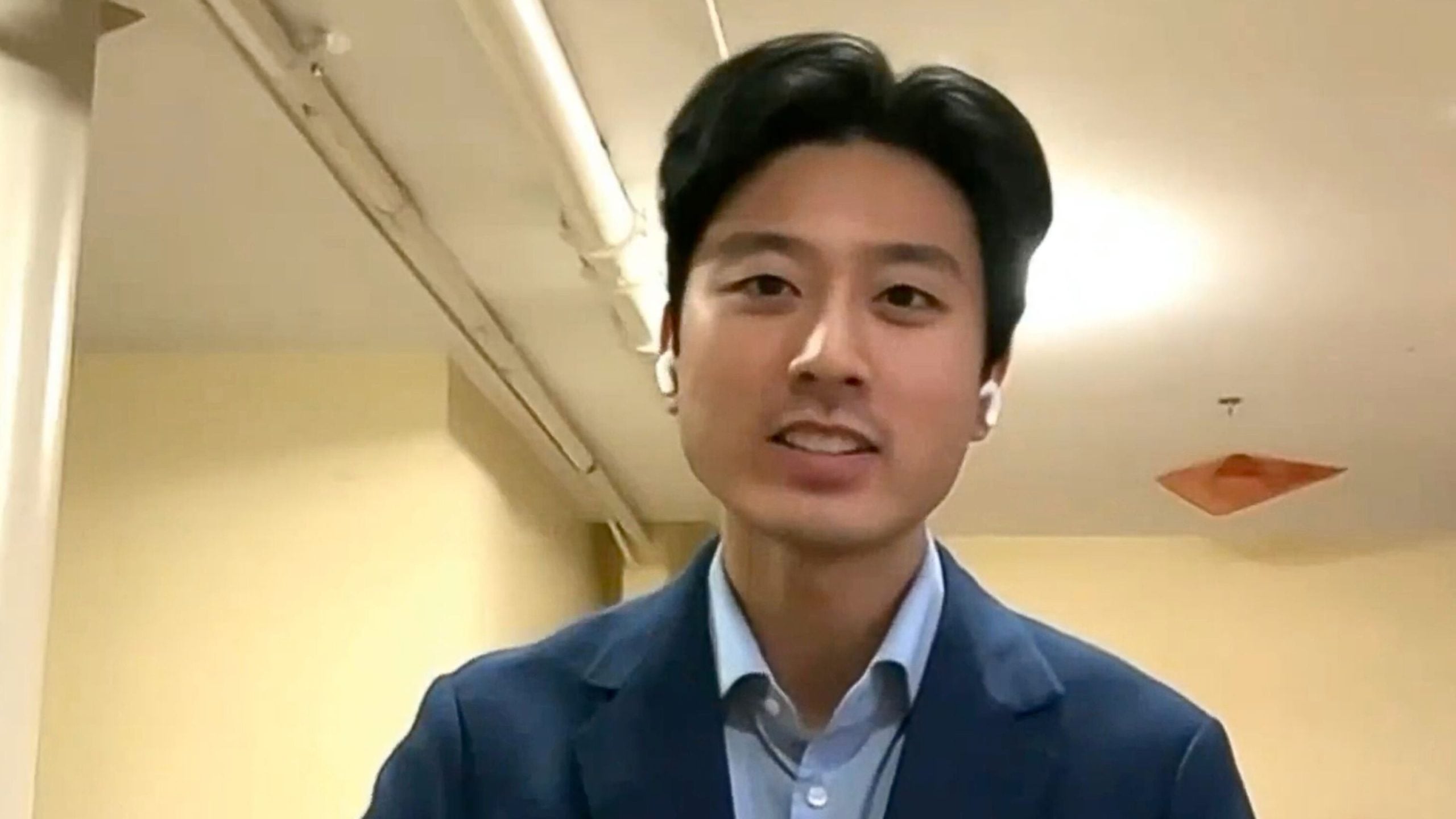 Alex Shieh wearing a blue suit and a shirt, with white earbuds, is speaking in an indoor setting.