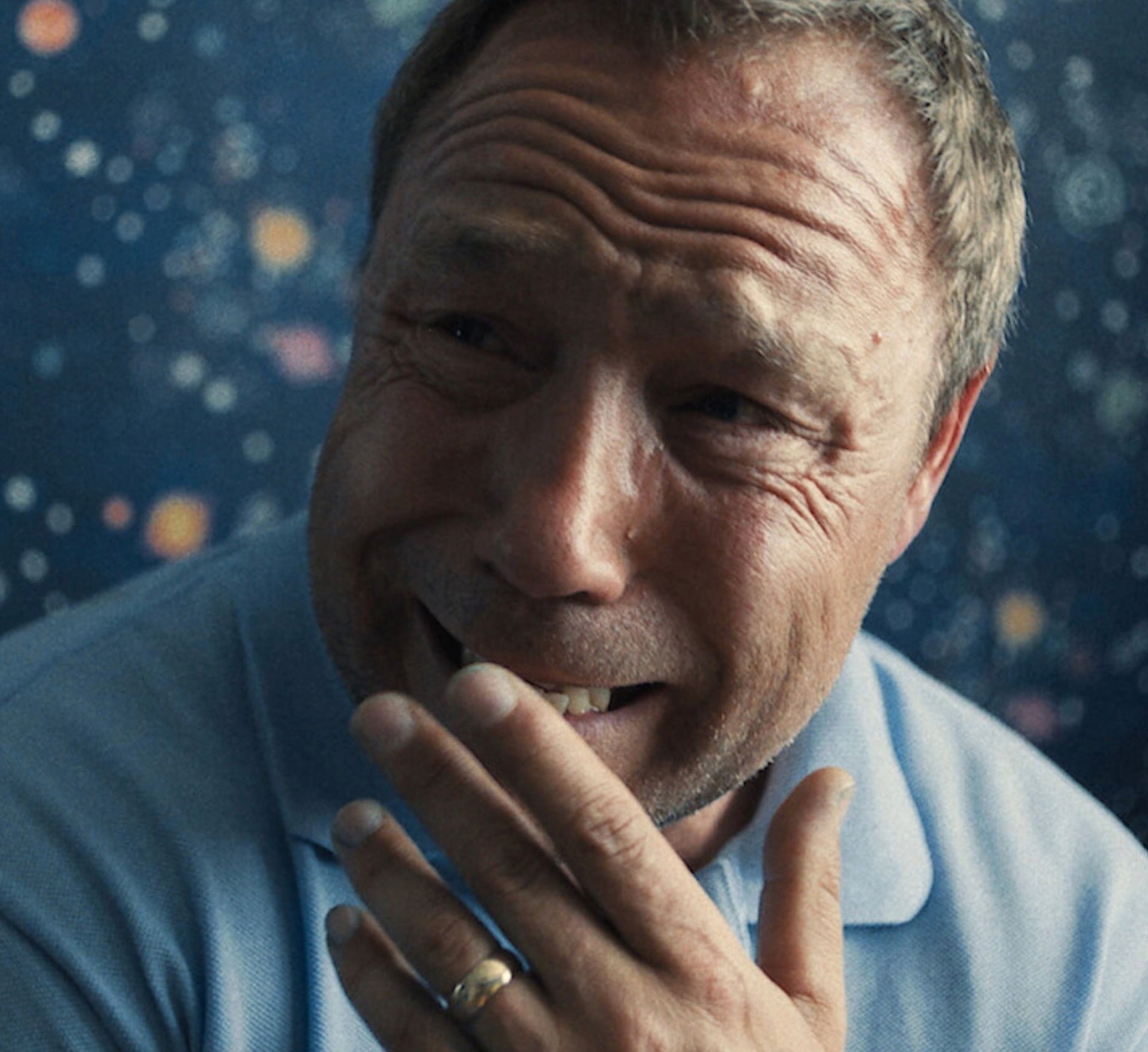 Steven Graham in a blue shirt laughing with his hand covering his mouth, against a starry, cosmic backdrop.