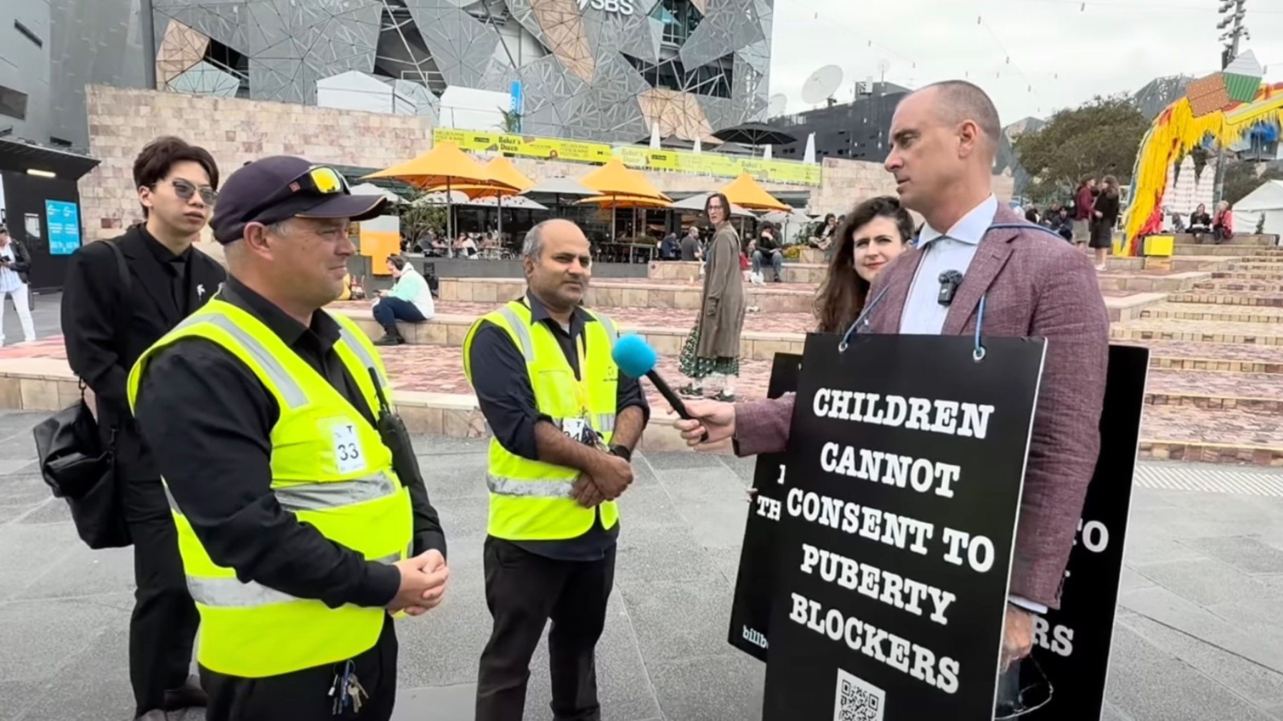 A person wearing a sign that reads "Children cannot consent to puberty blockers" is being interviewed by someone holding a blue microphone. Two men in yellow reflective vests stand nearby. The background features modern architecture and people sitting or walking in the public area.