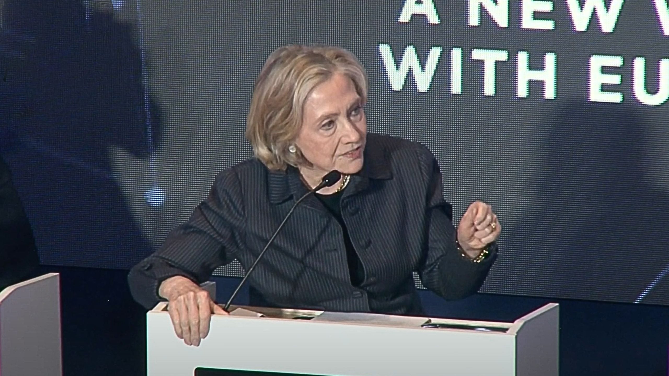 Clinton speaking at a podium with a microphone, dressed in a dark jacket, in front of a large screen displaying partial text.