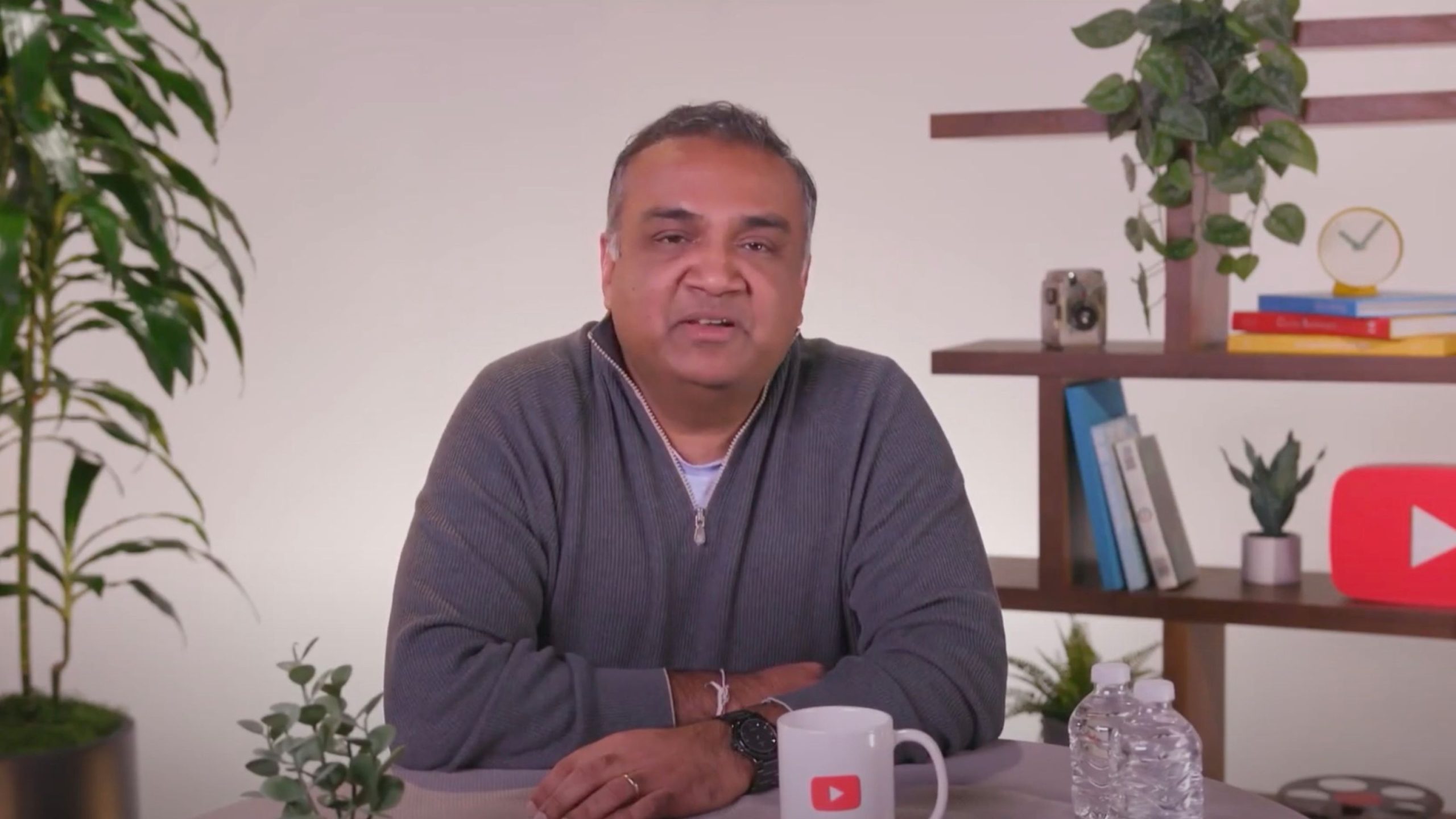 Mohan sitting at a table with folded arms, surrounded by potted plants and shelves with books and a clock in the background. The table features a YouTube-branded mug and two water bottles.