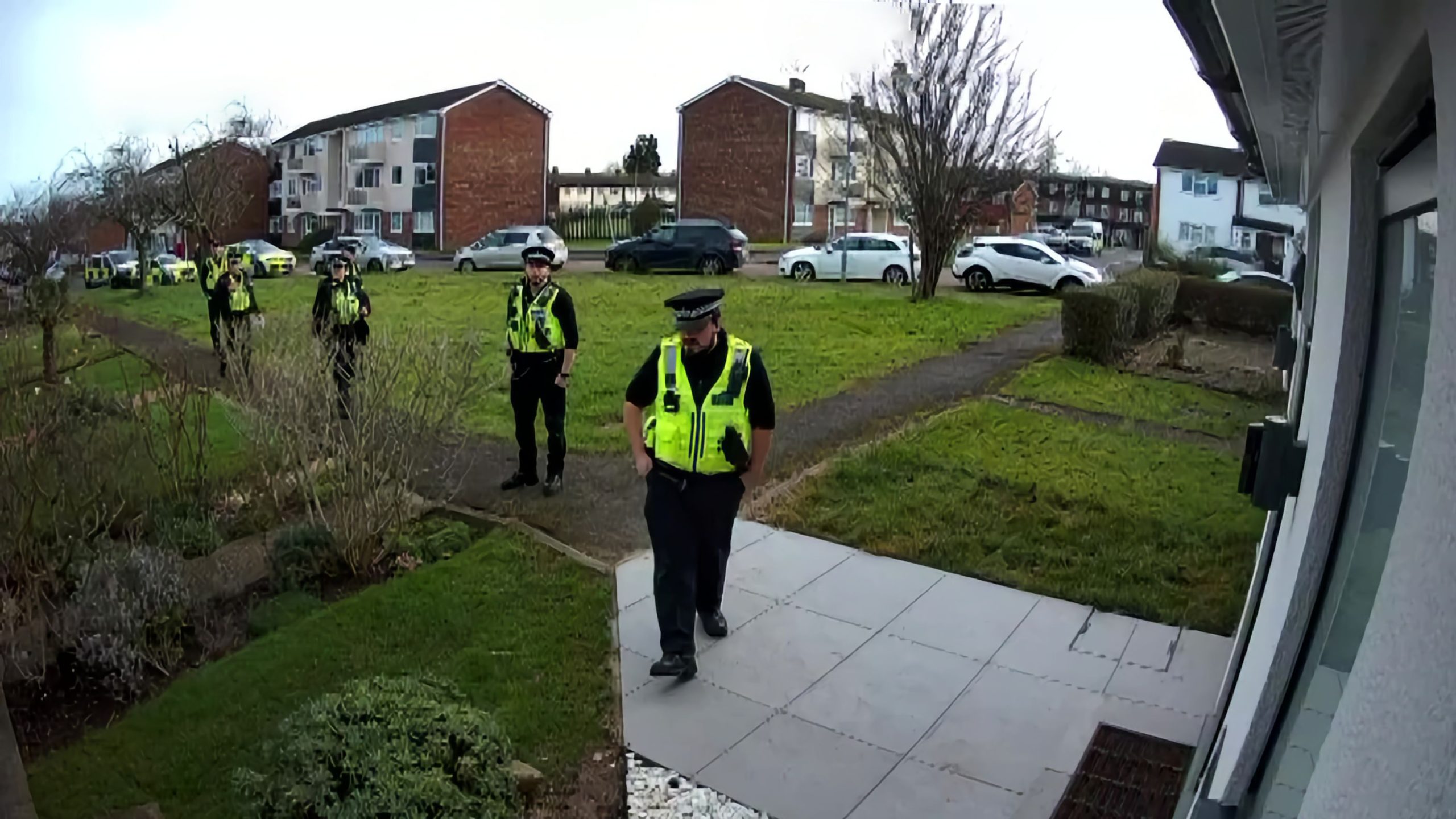 Uniformed police officers in high-visibility jackets approaching a residential doorstep in a suburban area, with apartment buildings and parked cars in the background.