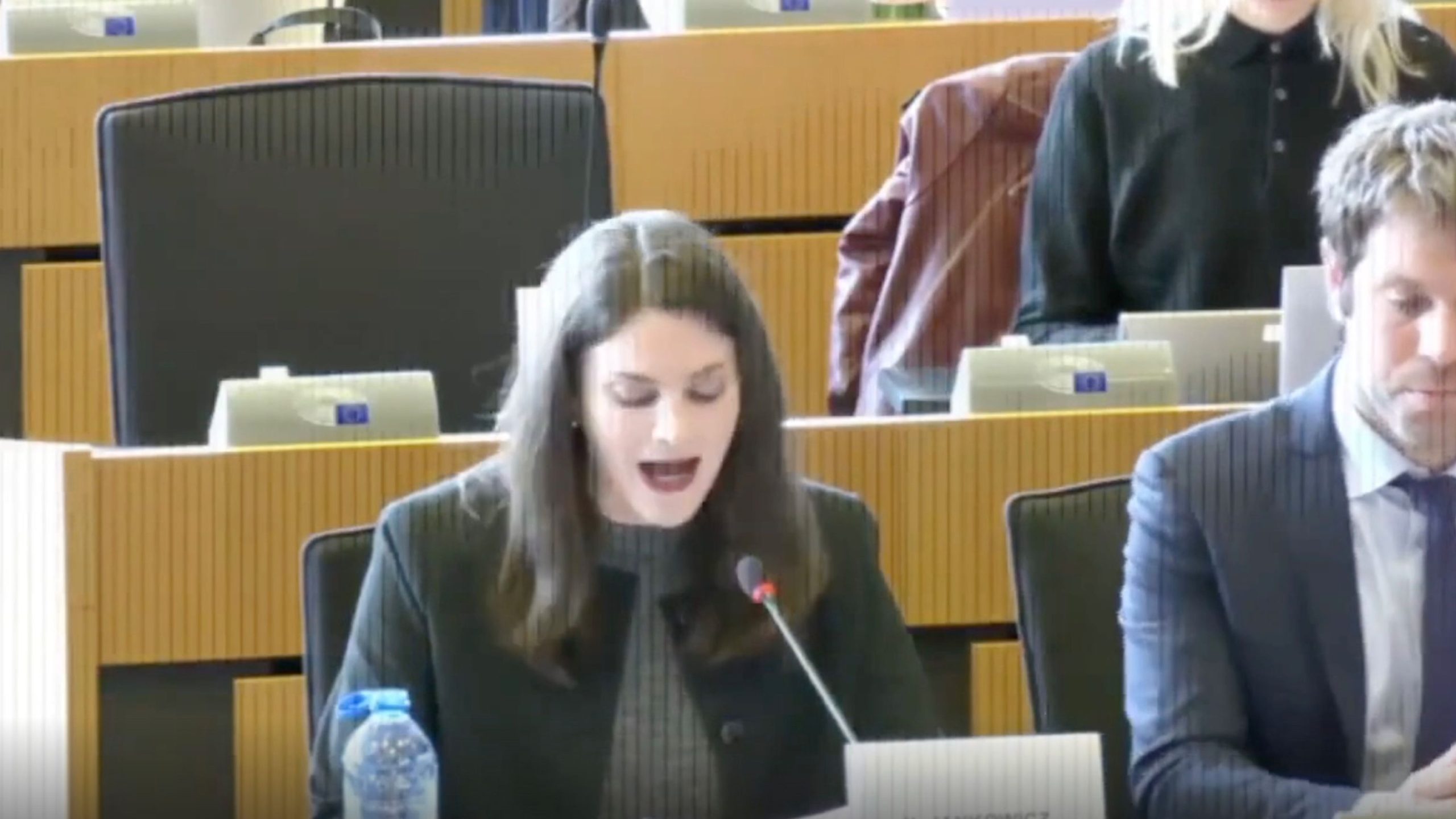 Jankowicz with long dark hair speaking into a microphone at a desk in a formal setting, with a man in a suit sitting to her right and another person visible in the background.