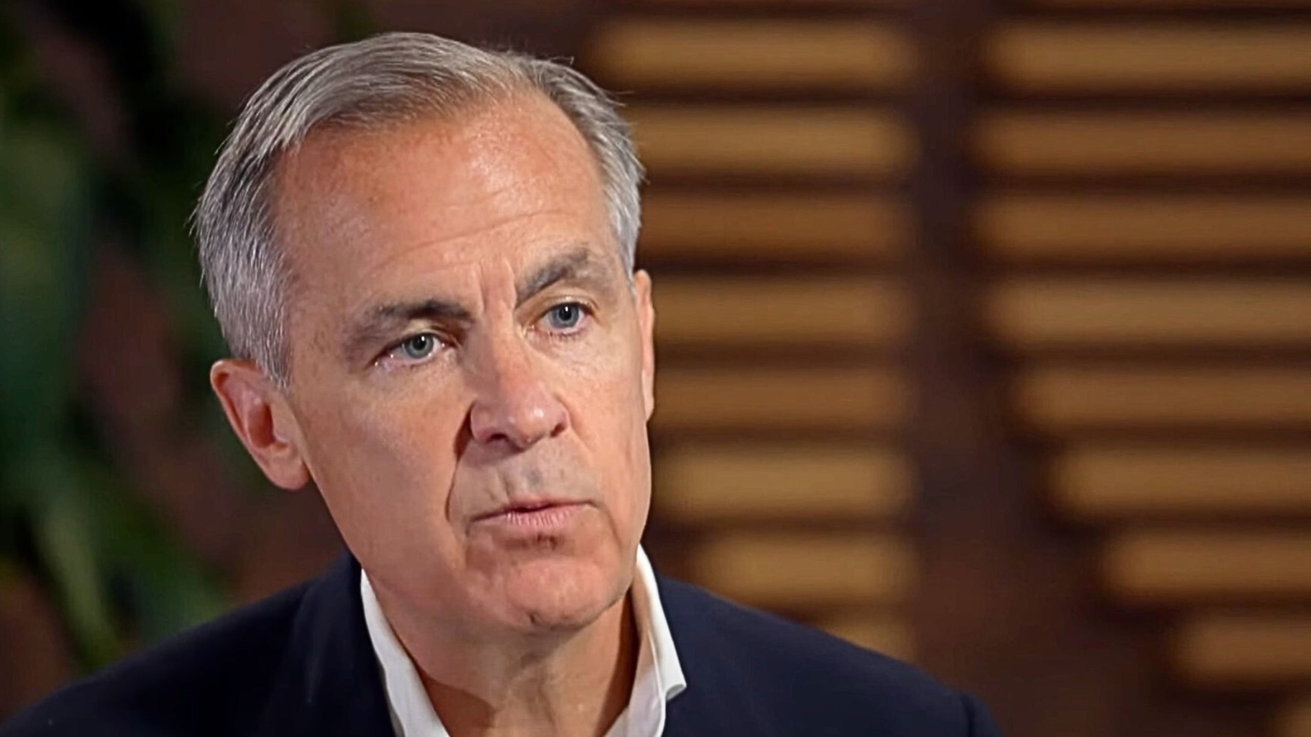 Carney with short gray hair wearing a dark jacket and white collared shirt, looking serious, with a blurred background.