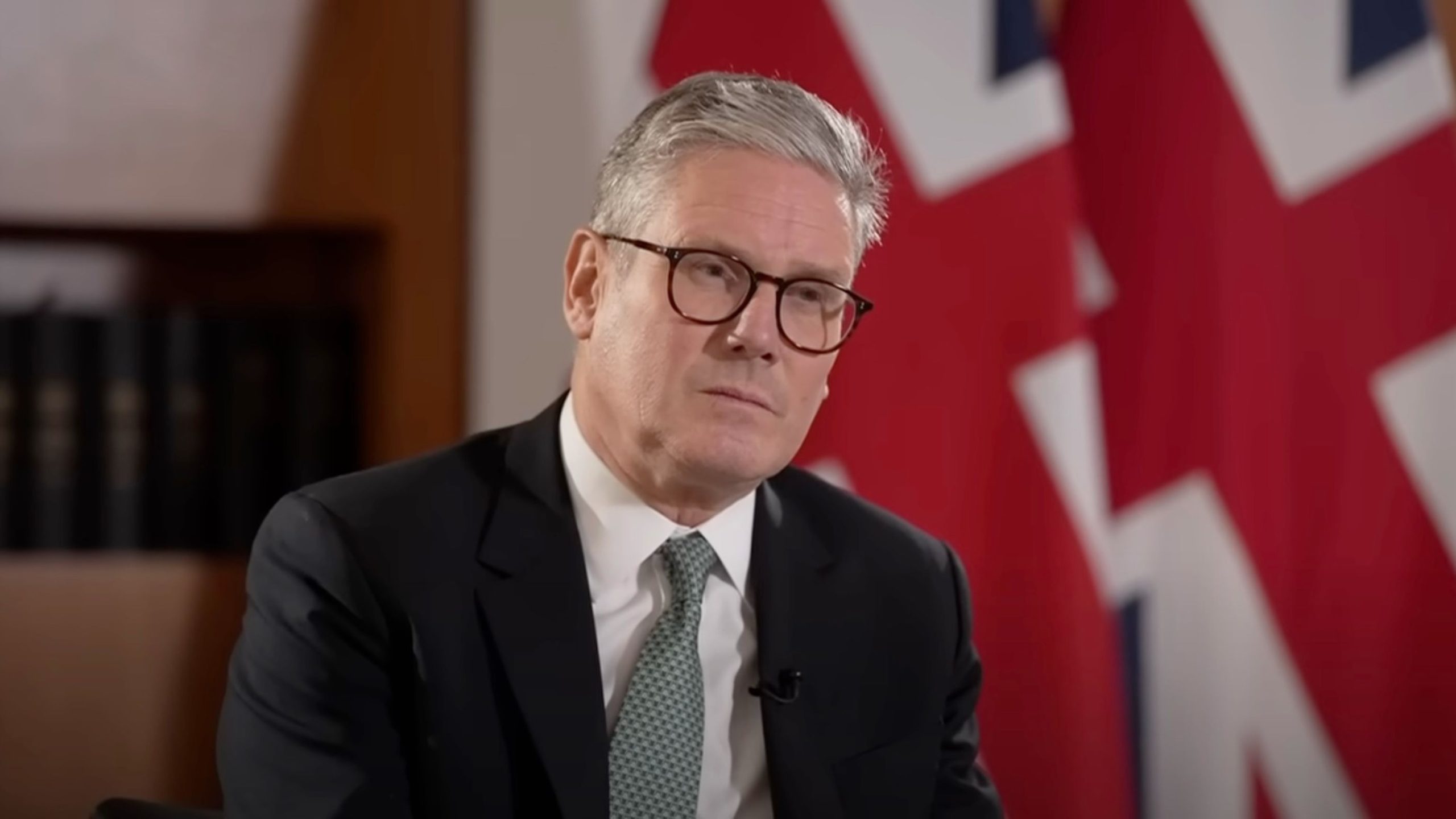 Starmer in a suit and glasses, sitting in front of a backdrop featuring parts of the UK flag, appears to be in an interview setting.