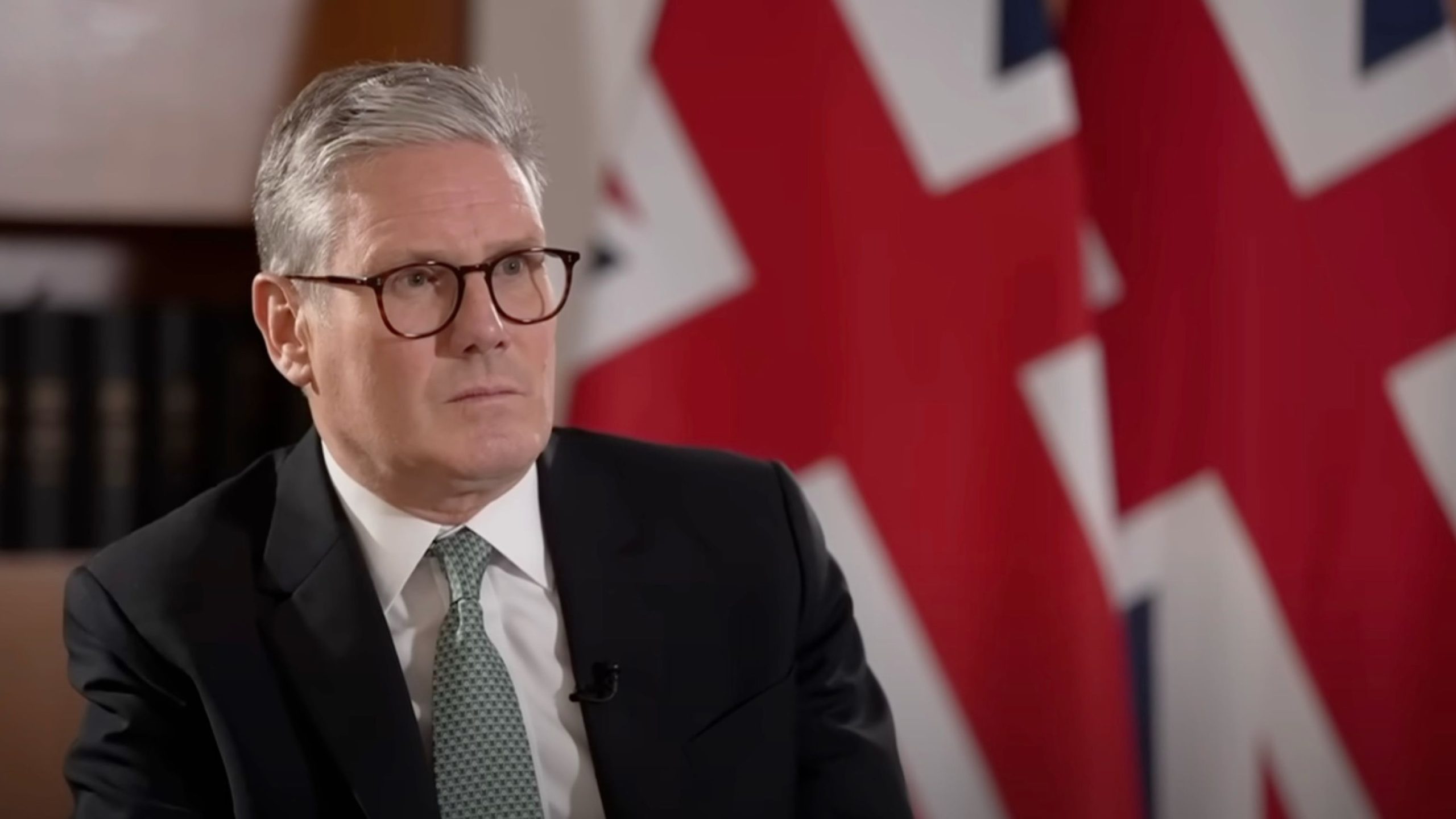 Starmer in a suit and glasses is seated in front of large Union Jack flags, appearing to be in a formal setting.