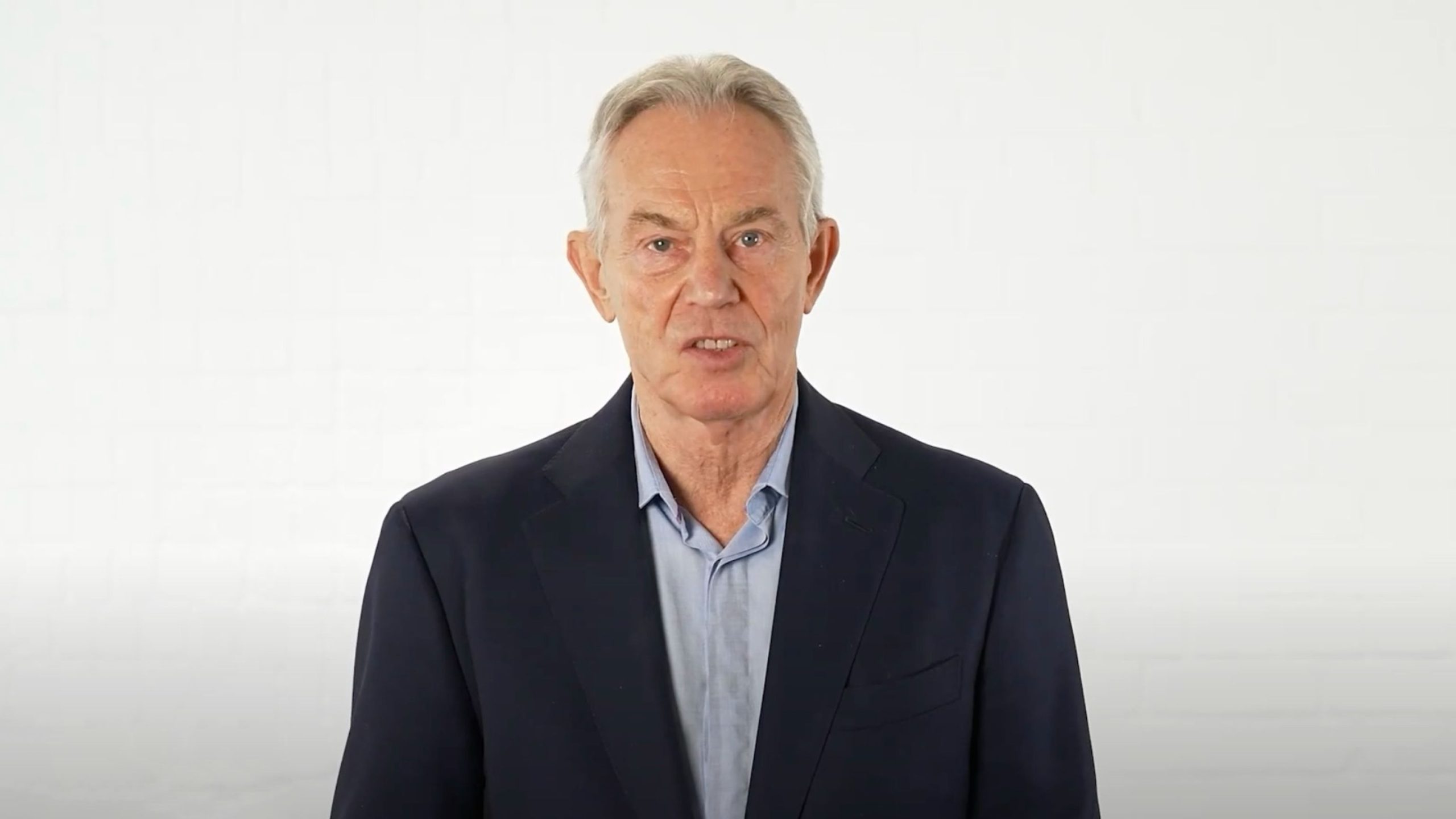 Tony Blair with gray hair wearing a dark blazer and light blue shirt standing against a plain white background