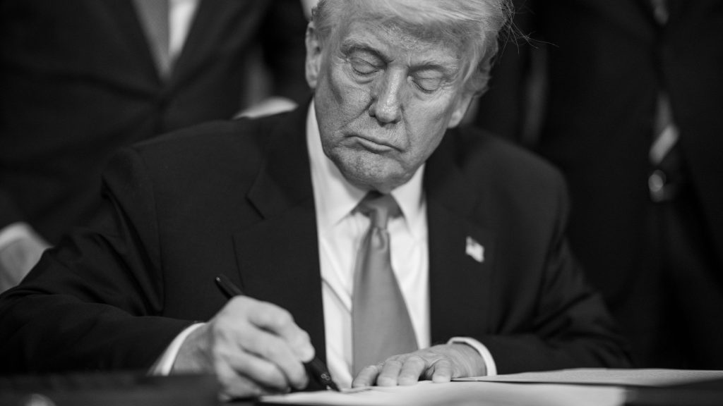 Trump in a suit and tie is signing a document with a pen, in a black and white photograph.
