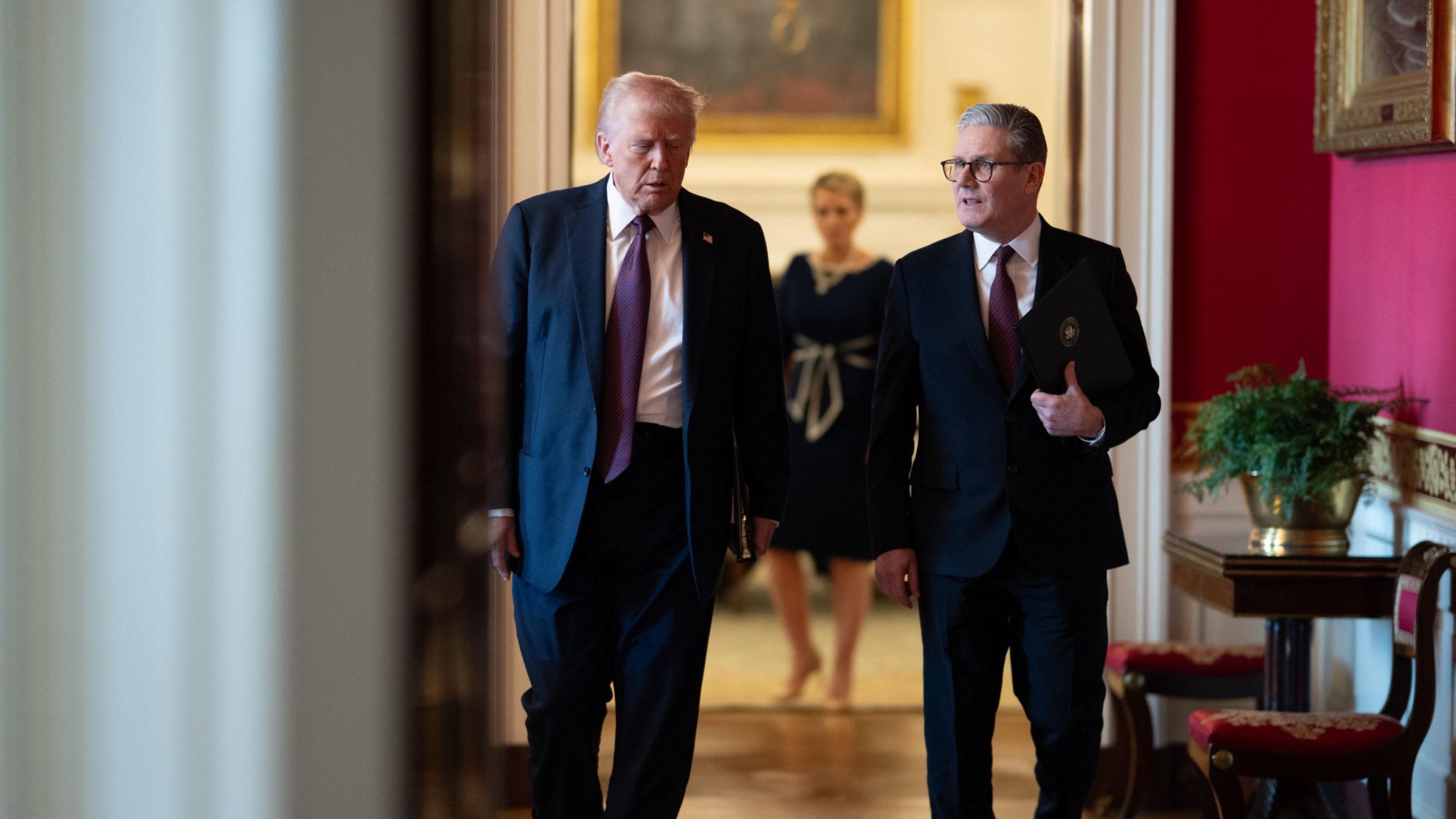 Trump and Starmer in suits walk down a hallway lined with red walls and ornate decor, engaged in conversation, with a woman in a black dress following behind them.