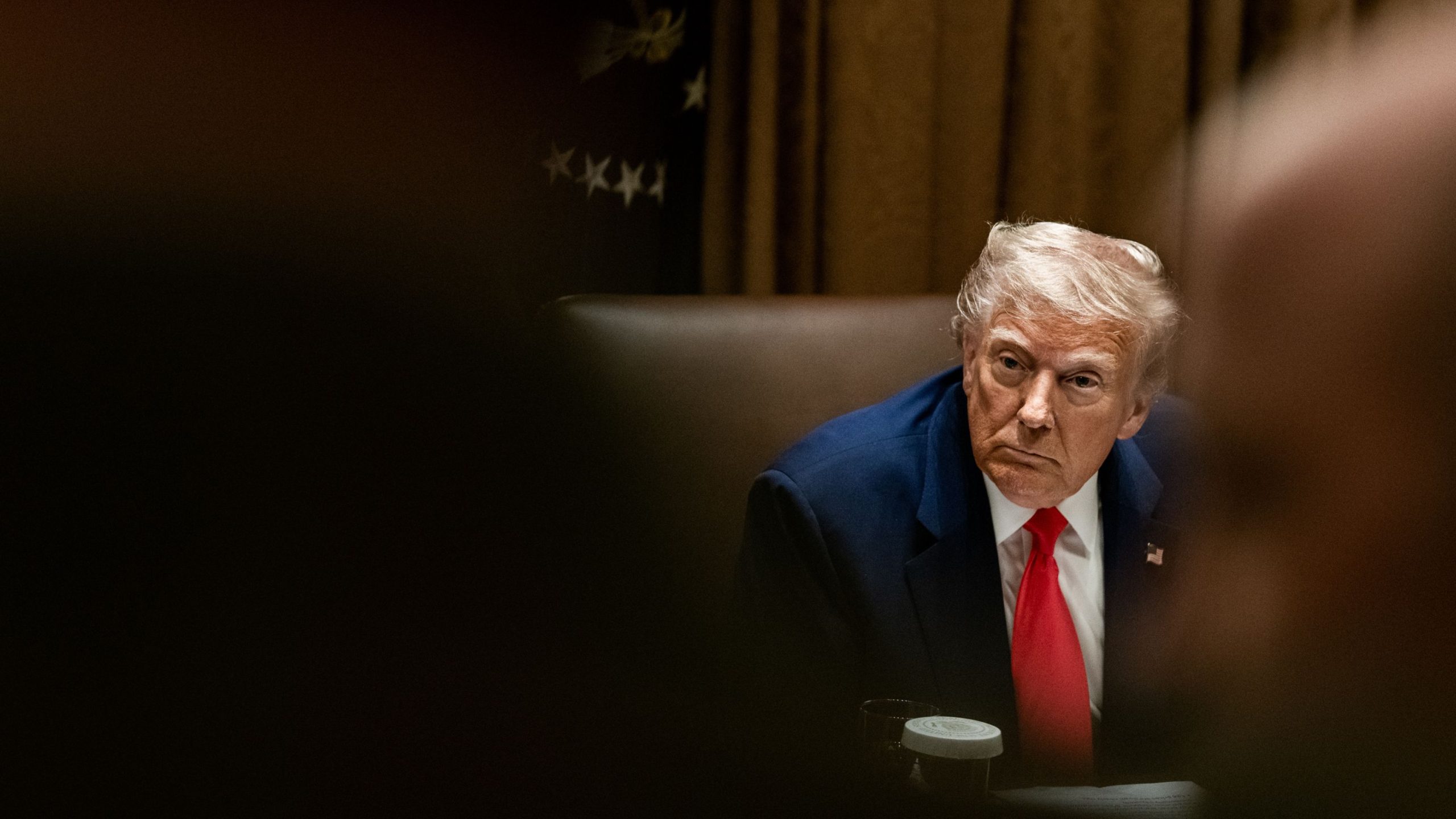 Trump in a dark suit and red tie sitting at a table, looking to his left with a serious expression, against a backdrop of dim lighting and partially visible stars on fabric.