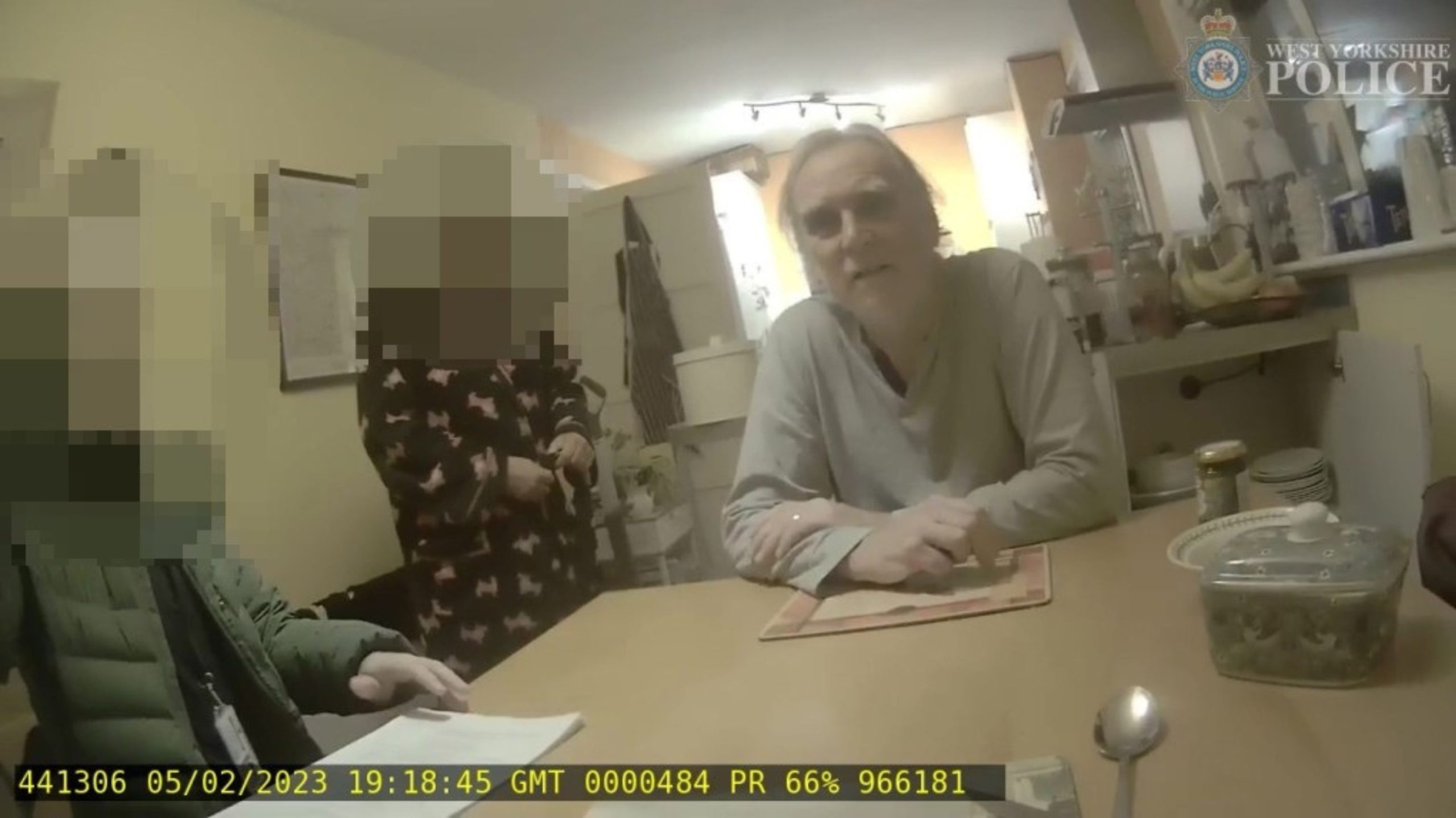 A person sitting at a kitchen table looks towards the camera, with two individuals blurred for anonymity. The setting is a domestic kitchen with visible cabinets, a banana bunch, and various kitchen items on a countertop. A timestamp and text overlay are present in the image.