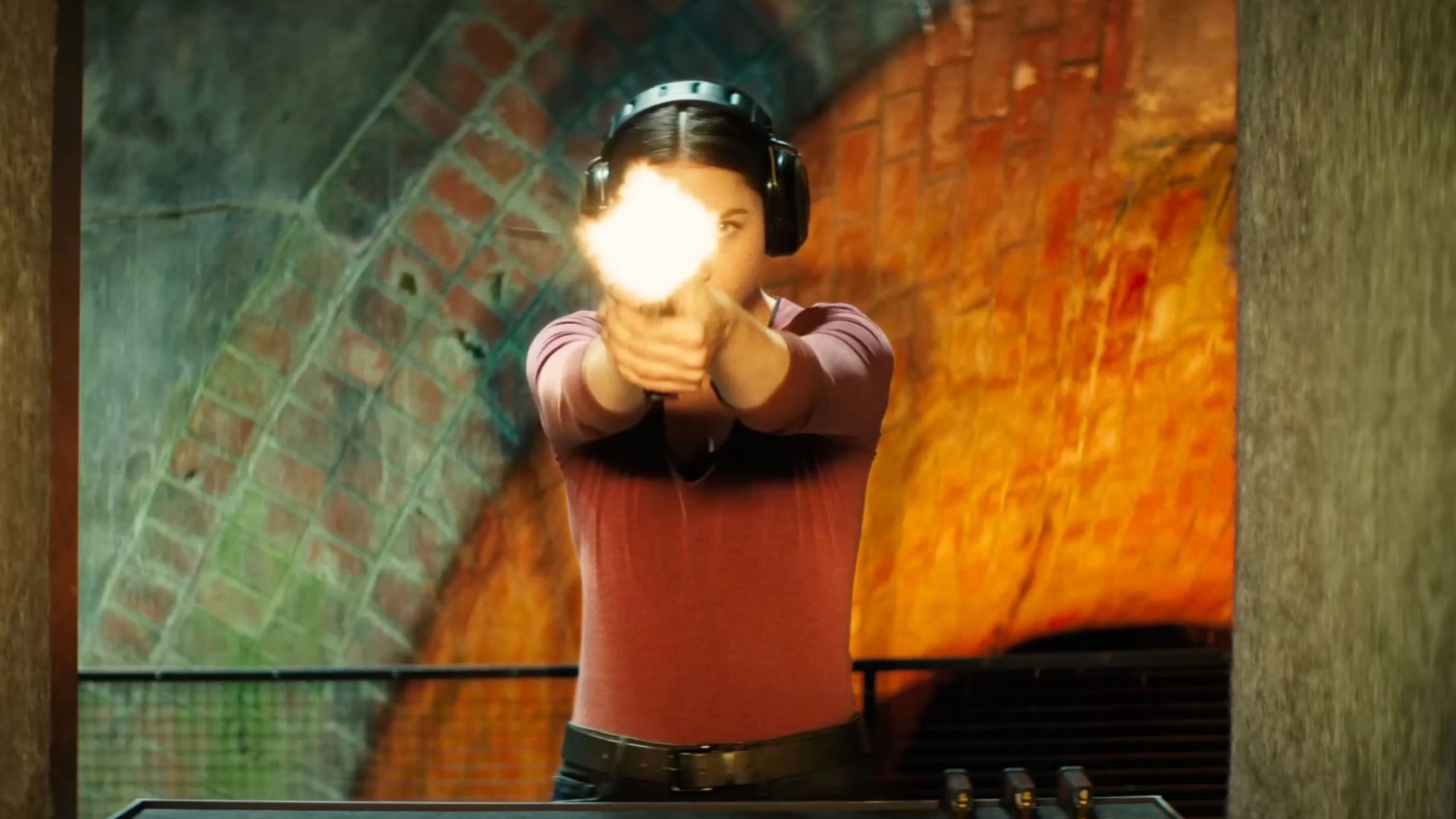 Ana de Armas wearing earmuffs firing a handgun at an indoor shooting range with a brick arch wall in the background.