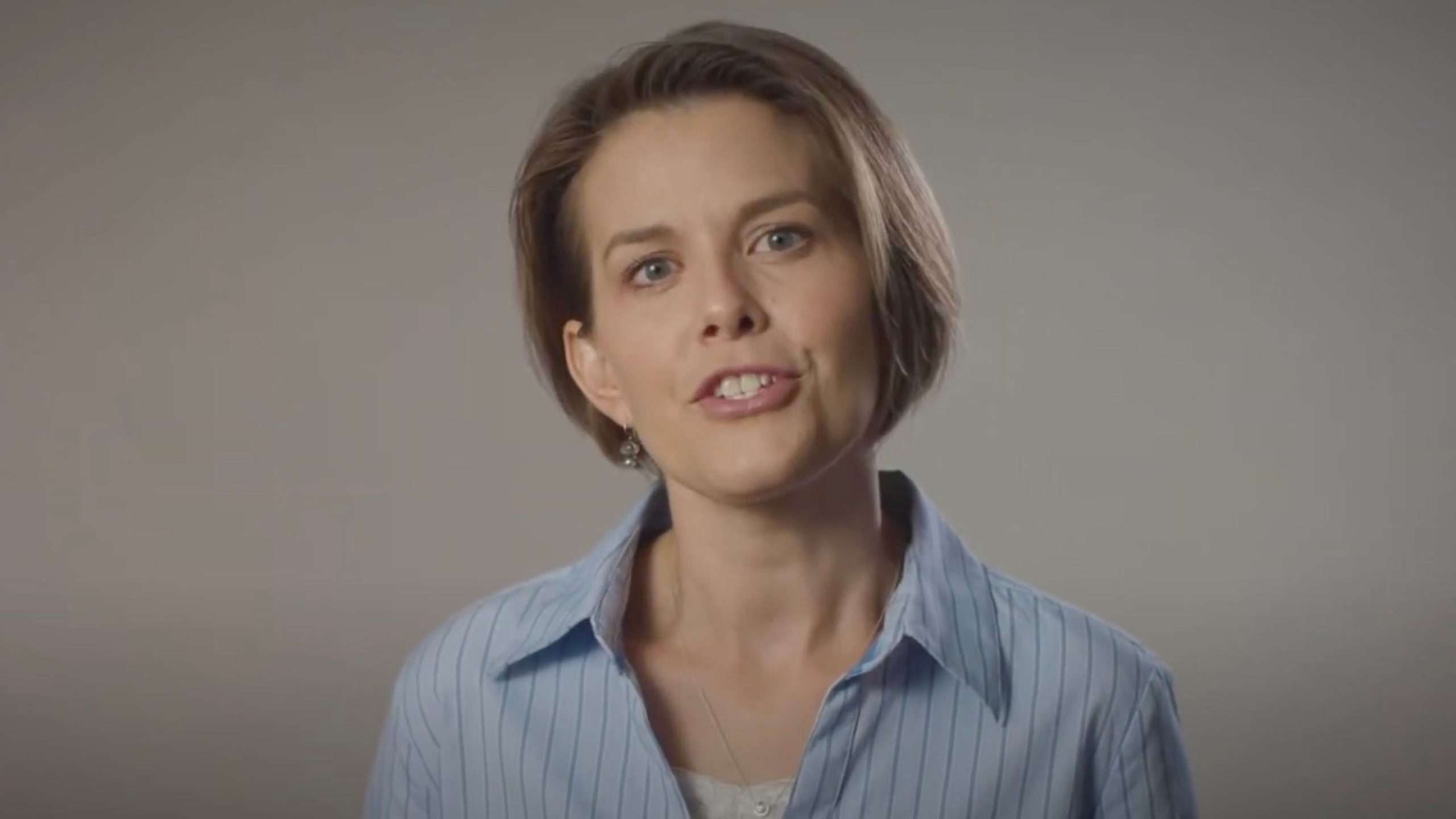 Woman with short brown hair wearing a light blue striped shirt speaking against a plain gray background.