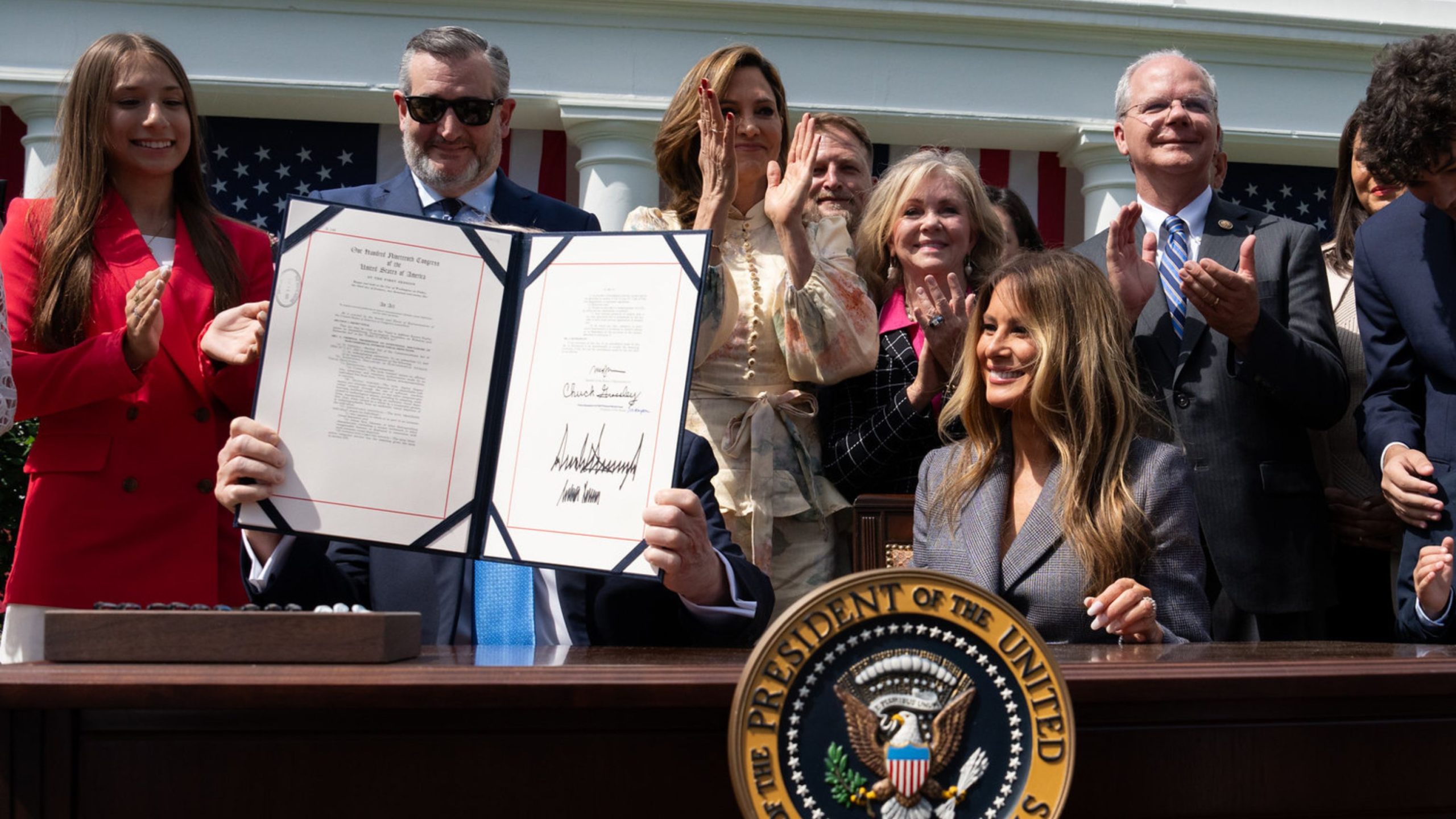 Trump seated at a desk holding up a signed document in front of his face while smiling people stand behind him applauding outside a government building with American flags and presidential seal on the desk.