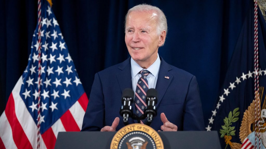 Joe Biden standing at a podium with the presidential seal, speaking with two microphones, with American flags in the background.
