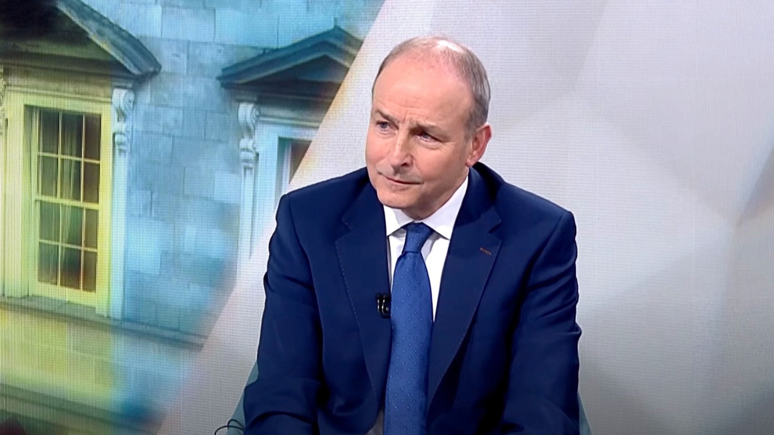 Micheál Martin in a dark blue suit and blue tie sitting and looking slightly to the side against a blurred backdrop of a windowed building facade and a light wall.