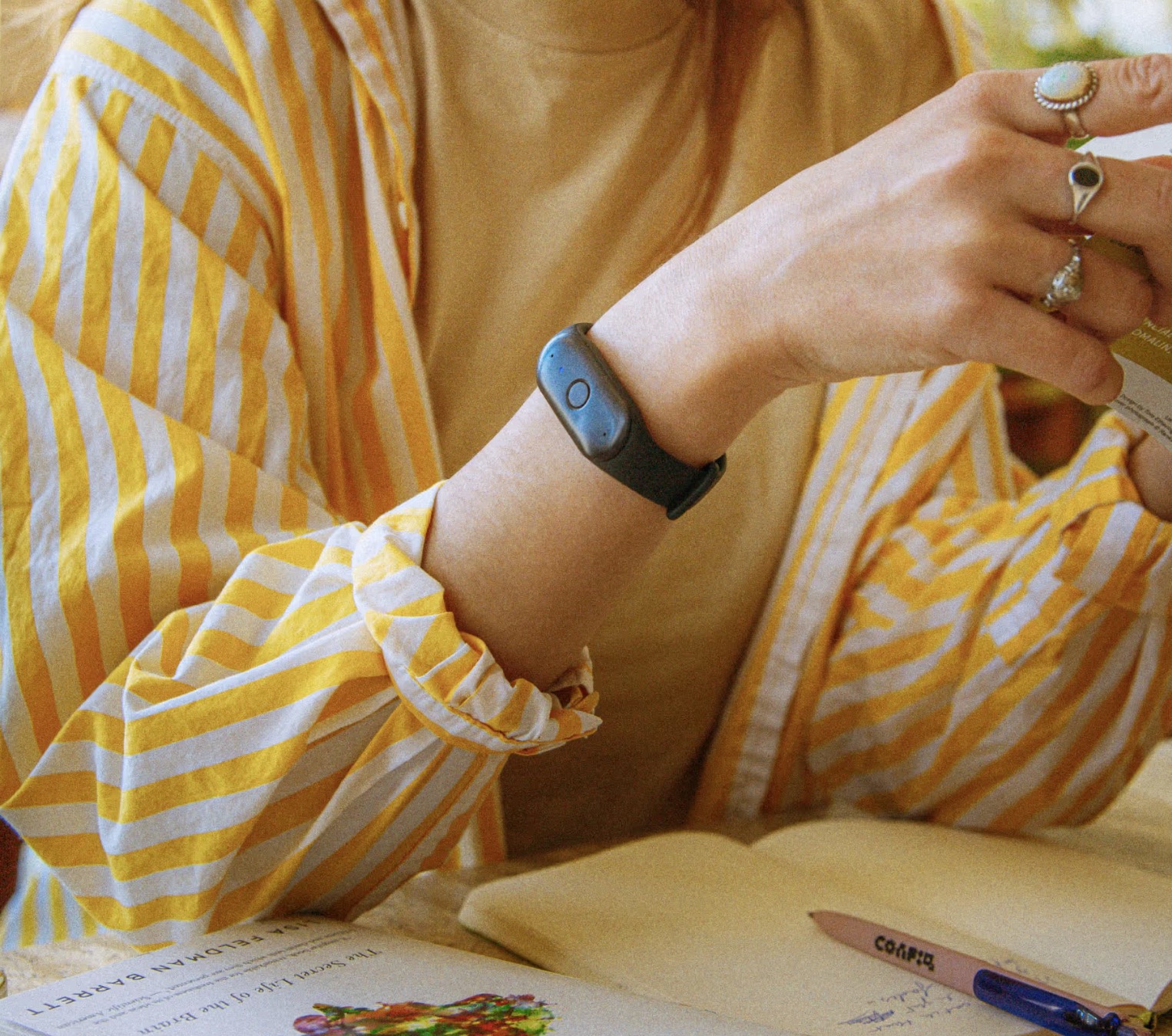 Person wearing a yellow and white striped shirt and a yellow top, with a black fitness tracker on their wrist and multiple rings on their fingers, holding a card or small booklet above an open notebook with a pen.