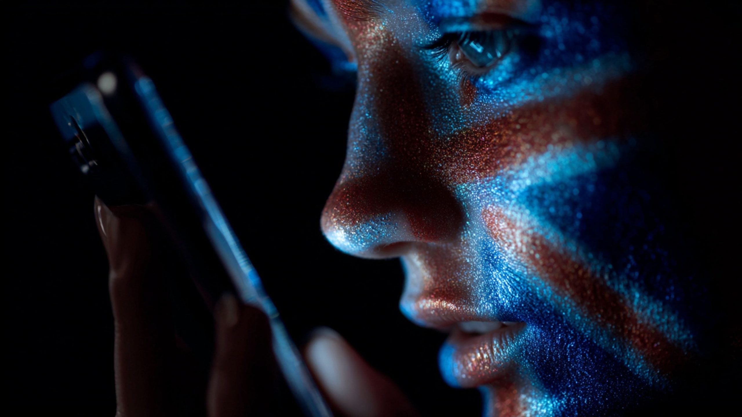 Close-up of a person's face with blue and red glitter paint reflecting light, looking intently at a smartphone held near their mouth against a dark background