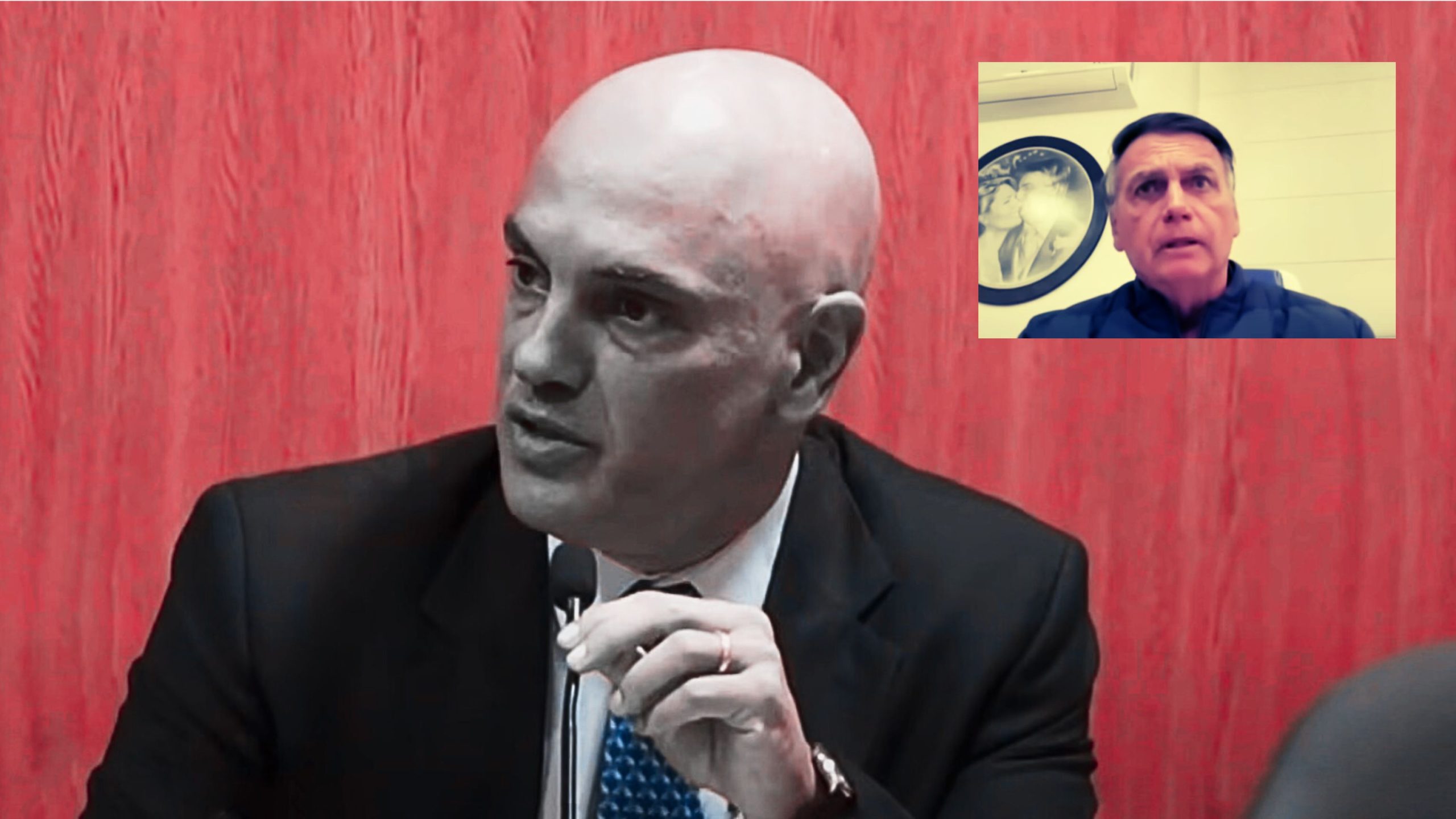 Moraes in a black suit and blue patterned tie speaking into a microphone against a red wooden background, with an inset showing another man in a dark jacket sitting indoors near a framed picture.