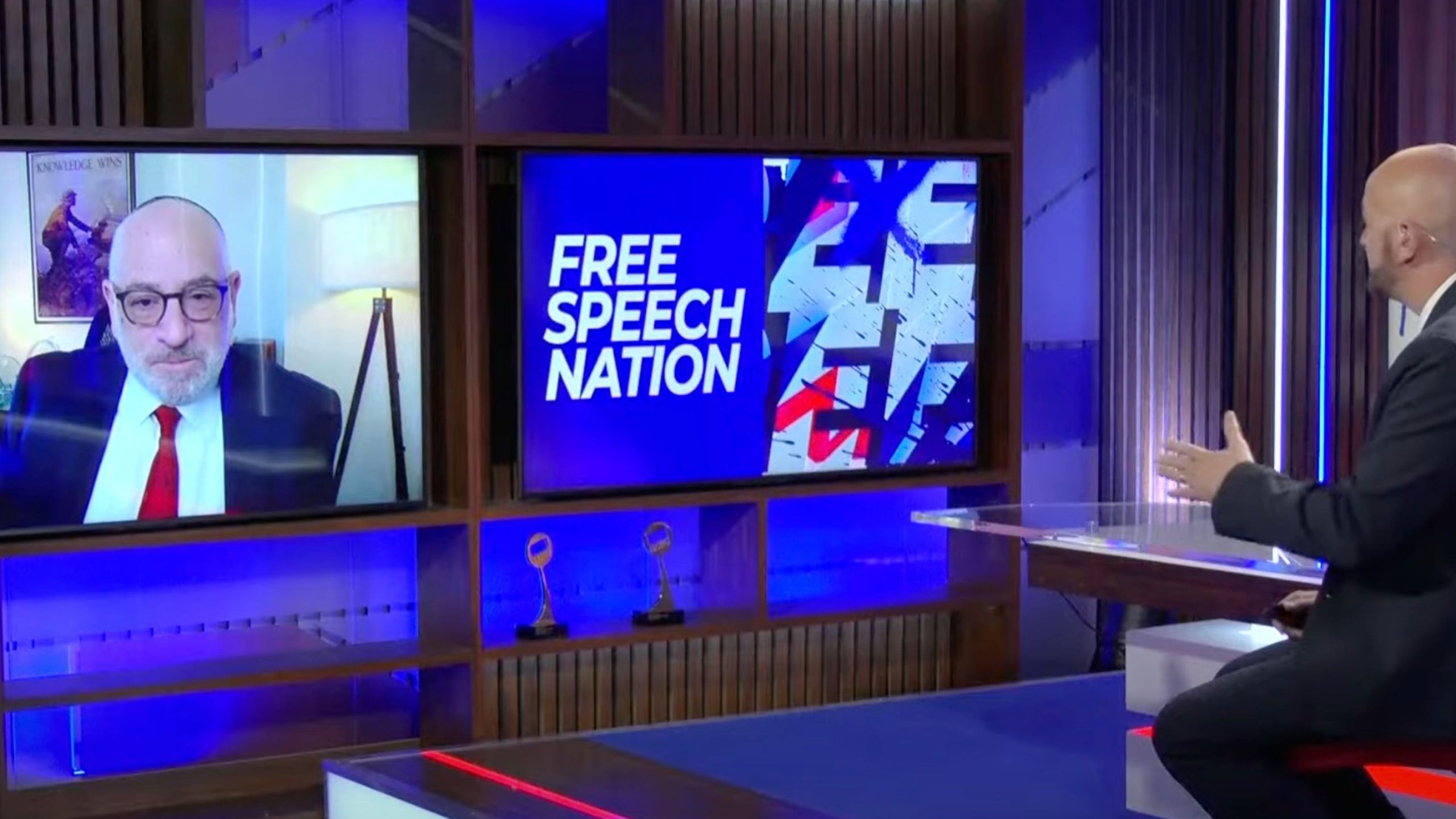 TV studio set with two large screens — left screen showing Coleman with glasses in a suit and red tie on a video call, center screen displaying the words FREE SPEECH NATION on a blue graphic, and at right Josh Howie in a suit gesturing from behind a clear desk.