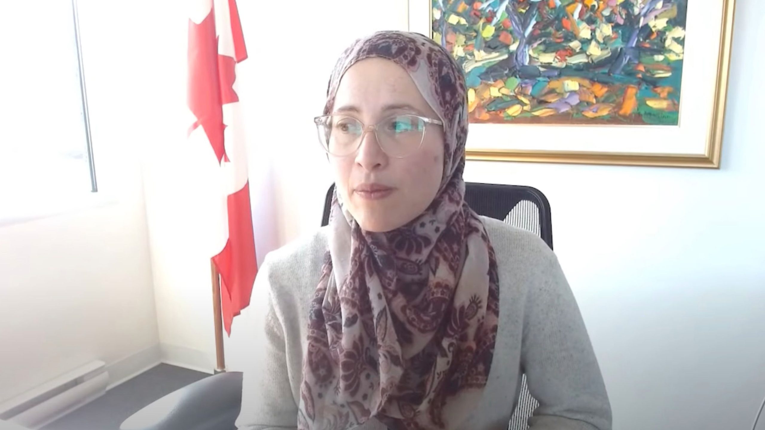 Amira Elghawaby wearing glasses and a patterned headscarf and a grey sweater seated in an office chair, with a Canadian flag to the left and a colorful framed painting on the wall behind.