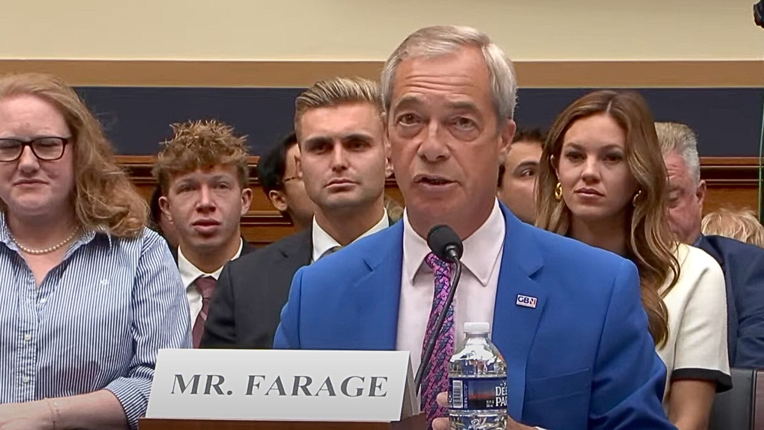 Nigel Farage in a bright blue suit and patterned tie speaking into a microphone at a formal hearing, seated behind a white name placard and a water bottle with several people seated behind him.