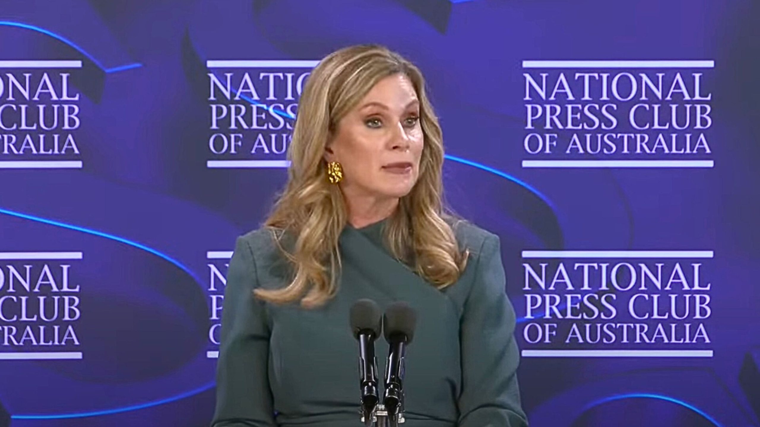Inman Grant in a teal dress and gold earrings speaking into two microphones at a podium, with a purple backdrop displaying repeated "National Press Club of Australia" logos.