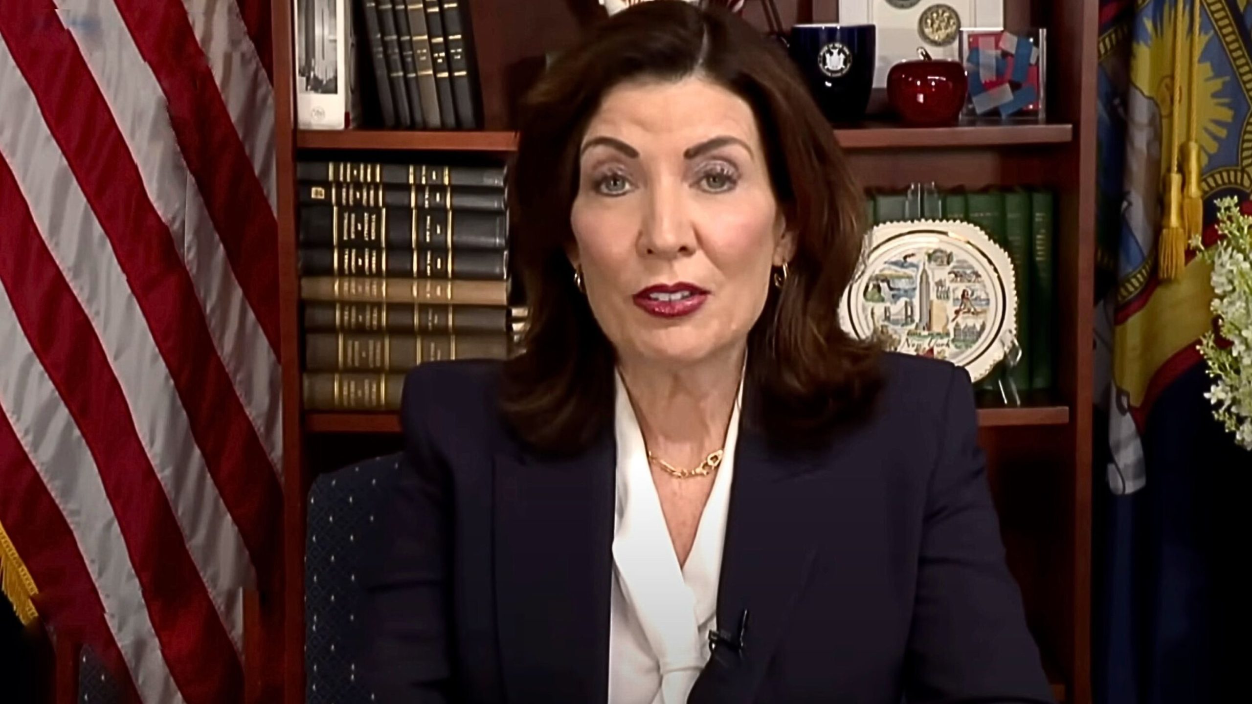 Hochul with shoulder-length dark hair wearing a navy blazer and white blouse speaking from an office, seated before a bookshelf filled with bound volumes, a decorative plate, and two flags including the U.S. flag.