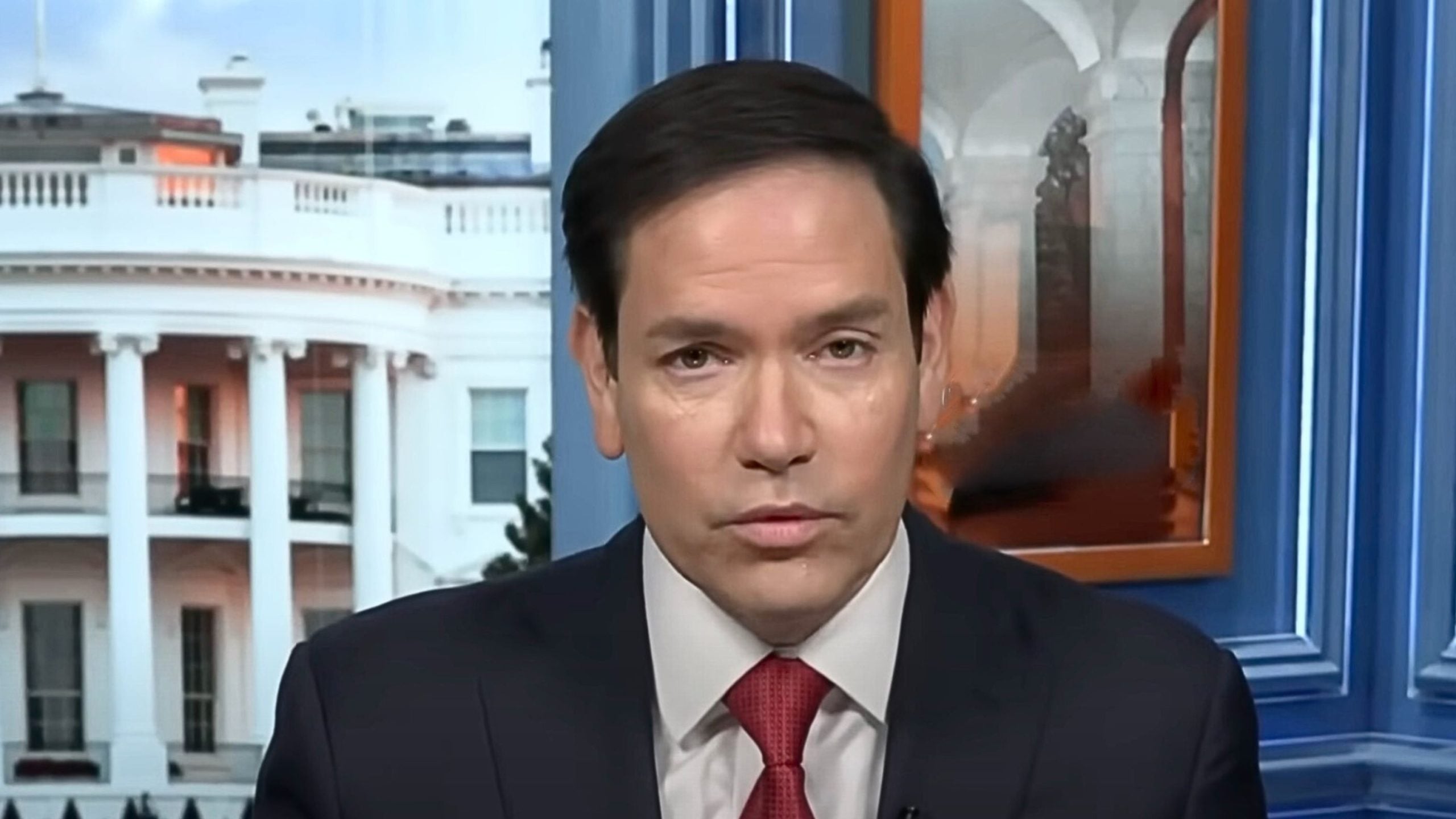 Close-up of Rubio with dark hair wearing a dark suit, white shirt and red tie speaking on camera in a studio with a view of the White House exterior through a window behind him.