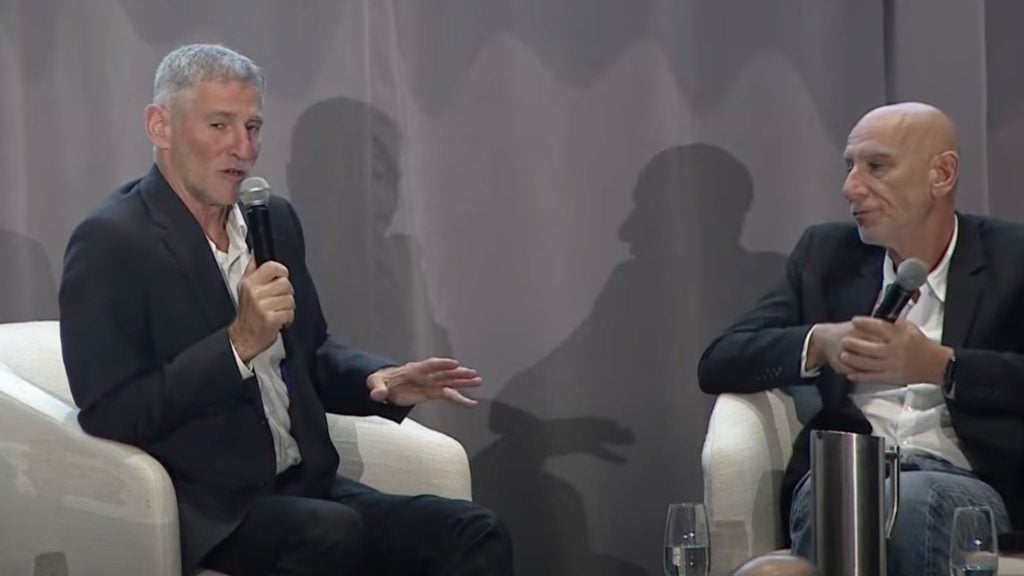 Golan and the host in dark blazers sit in white armchairs on a stage, each holding a microphone and gesturing while talking in front of a gray curtain that casts their shadows; a water pitcher and glasses sit on a small table beside them.