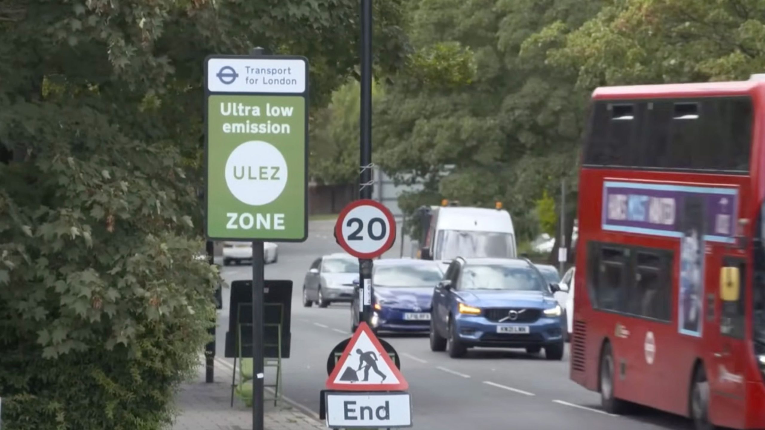 London street scene with a green Transport for London 'Ultra low emission ULEZ ZONE' sign, a circular 20 mph sign above a triangular roadworks sign reading 'End', and traffic including a blue car, white van and a red double-decker bus passing under trees.