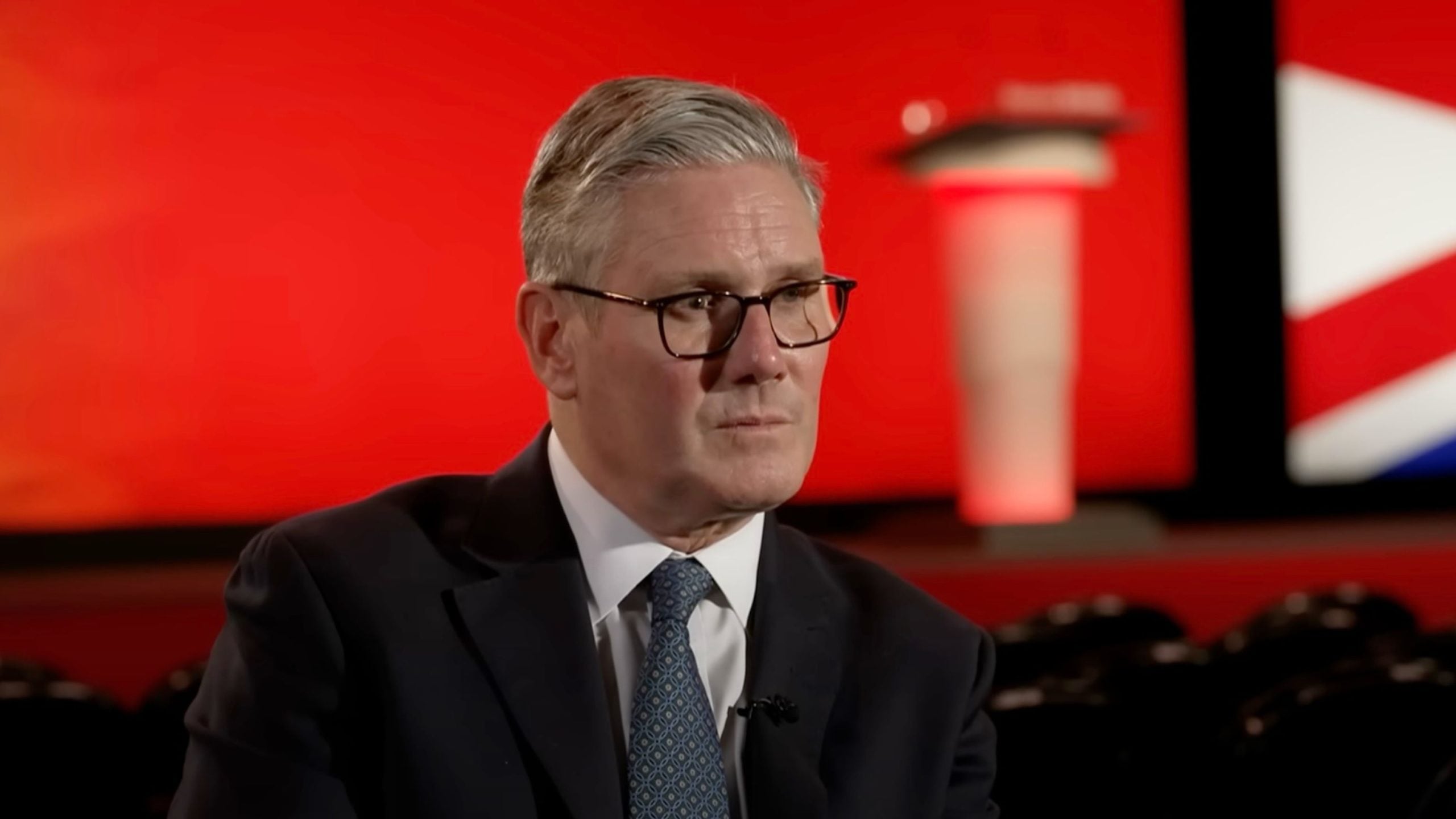 Starmer in a dark suit and patterned tie, seated and looking serious against a red stage backdrop with a blurred lectern and a fragment of a Union Jack flag.