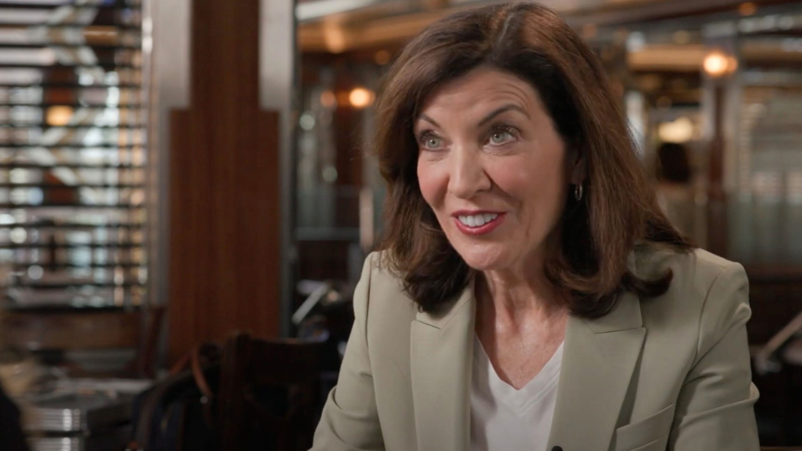 Hochul with shoulder-length brown hair smiling and speaking, wearing a pale blazer over a white top, seated in a warm wood-paneled restaurant with blinds and soft lights in the background.