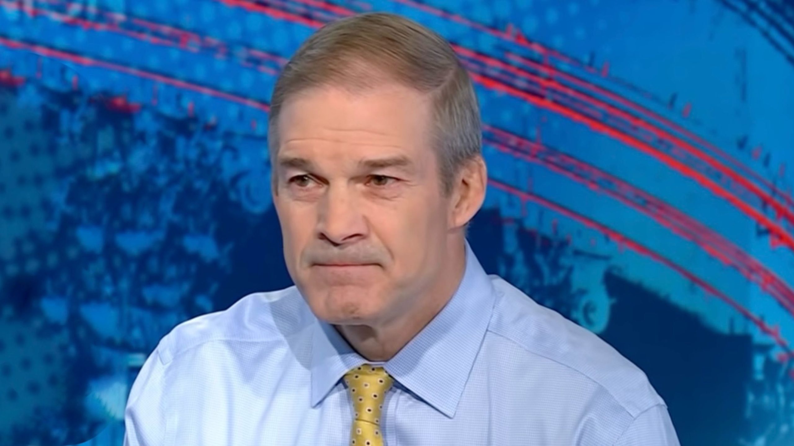 Jim Jordan in a light blue dress shirt and yellow patterned tie with a serious expression against a blue studio backdrop featuring red curved lines.
