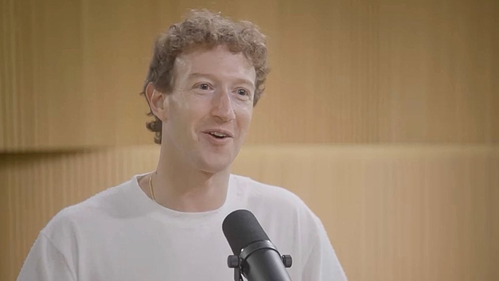 Zuckerberg with short curly light-brown hair wearing a white T-shirt, speaking into a black microphone in front of a wood‑paneled studio background.