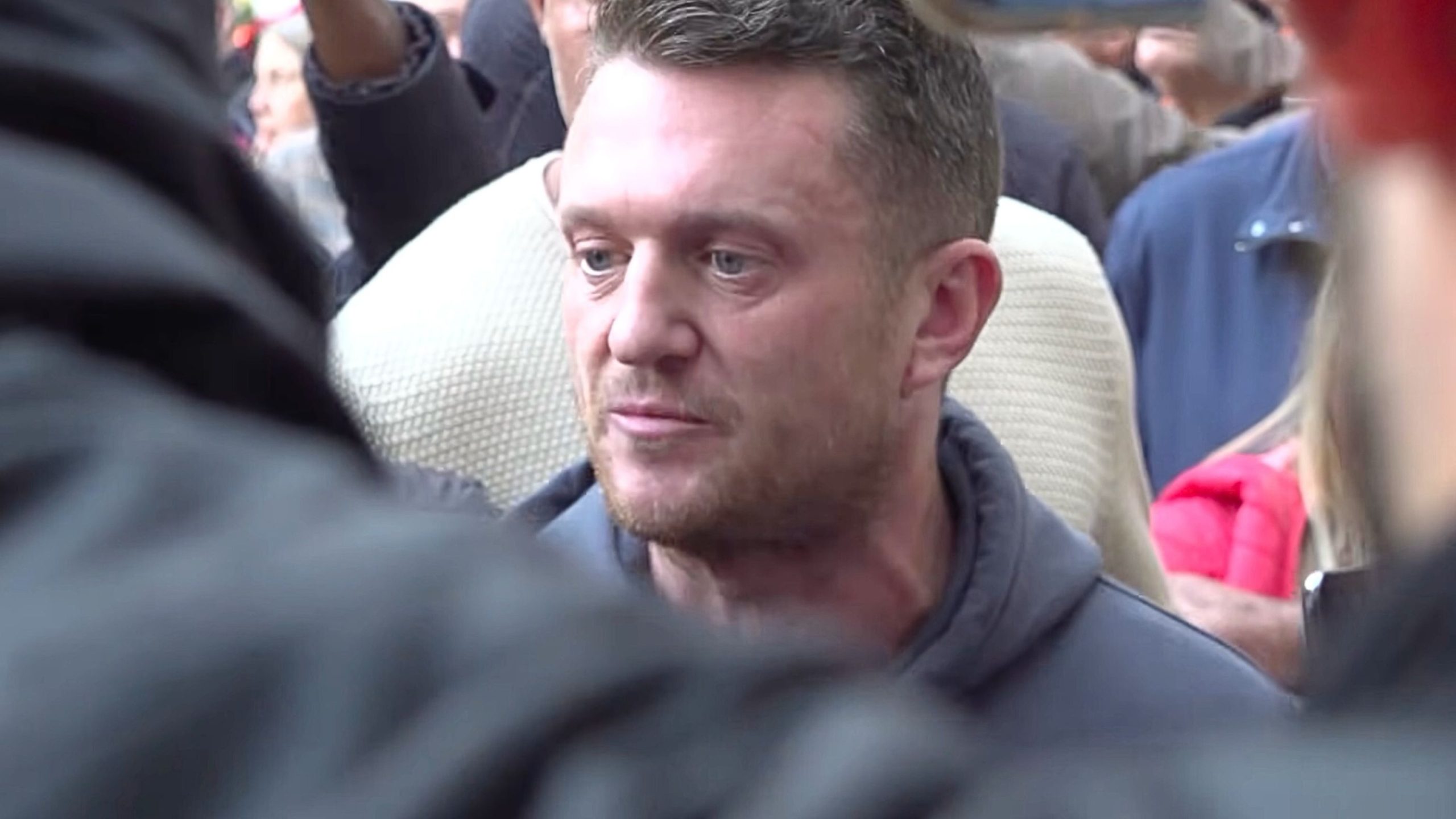 Close-up of a light-skinned man with short brown hair and stubble wearing a dark hoodie, looking slightly to the side with a serious expression while surrounded by a crowd of people.