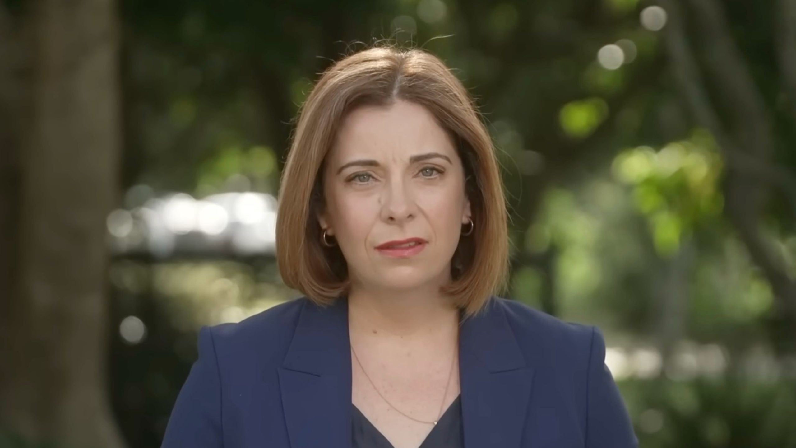 Anika Wells with shoulder-length brown hair wearing a navy blazer and small hoop earrings, looking serious outdoors.