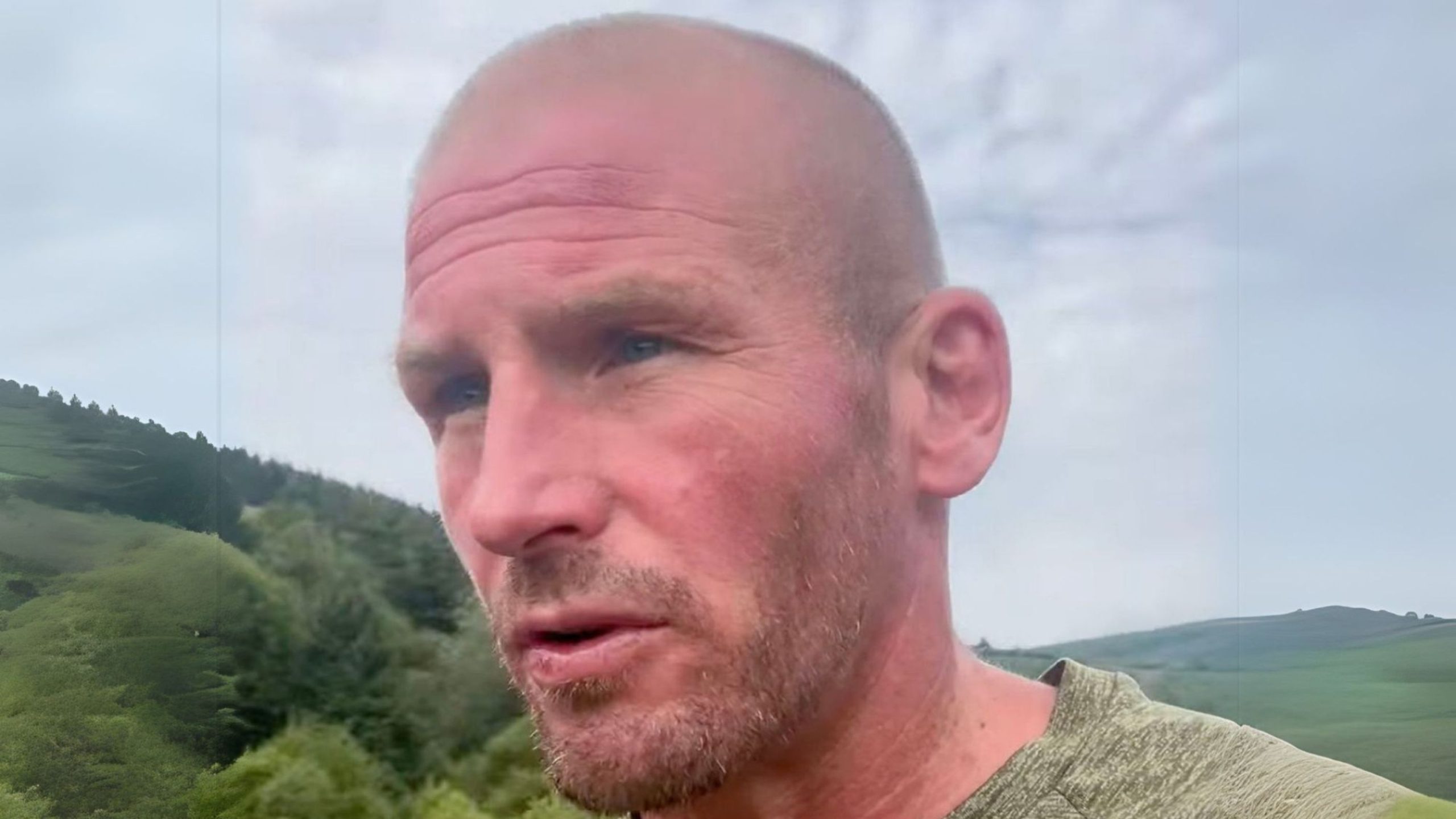 Jamie Michael with short stubble and blue eyes looking slightly off-camera, standing outdoors with rolling green hills and an overcast sky behind him.