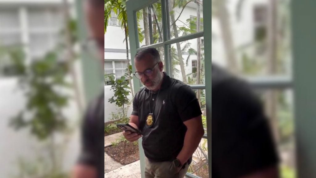 Cop wearing glasses and a badge on a lanyard reads his phone by a multi-pane window with plants outside.