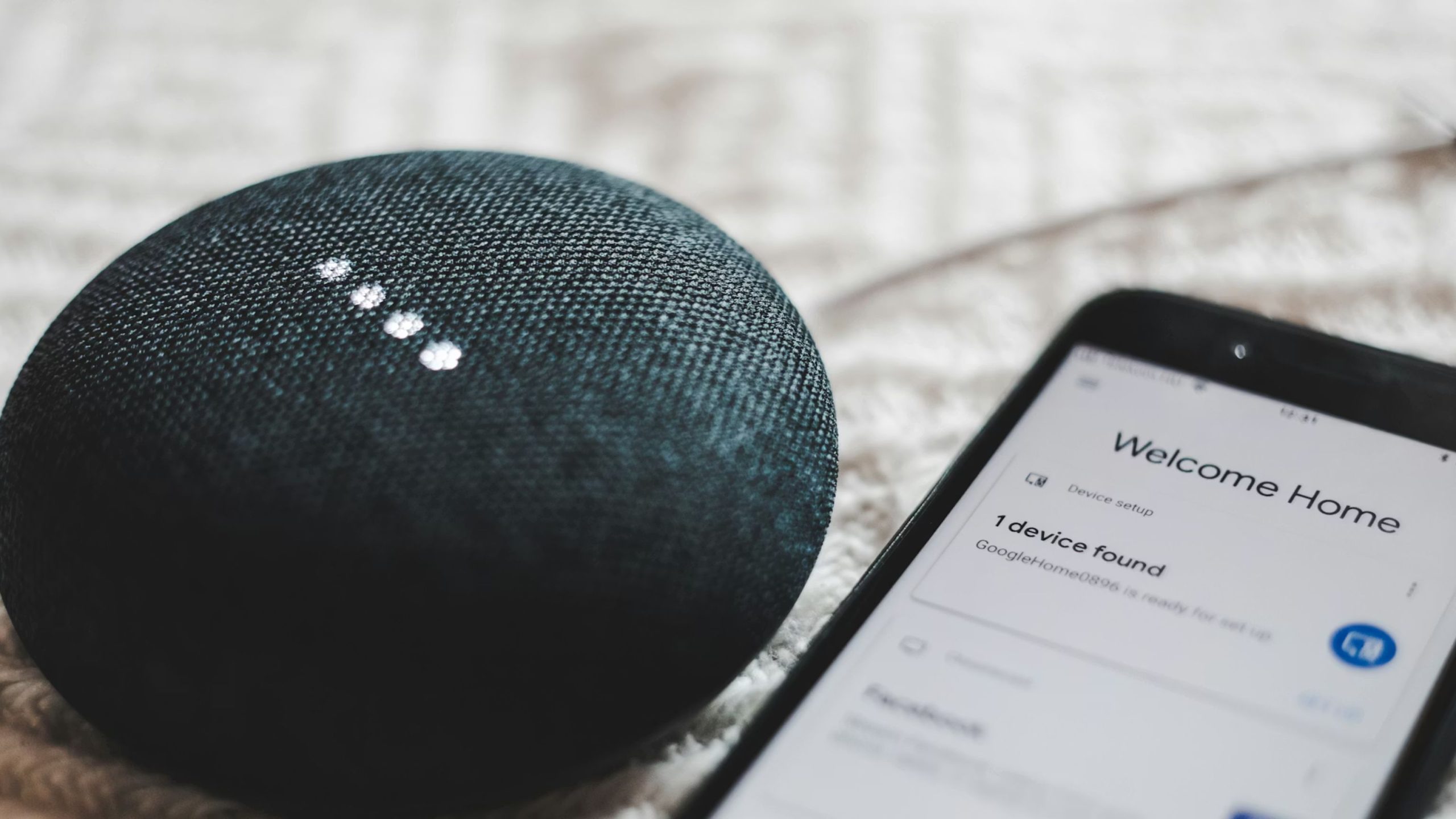 Round dark fabric smart speaker with four illuminated white dots next to a phone displaying Welcome Home device setup
