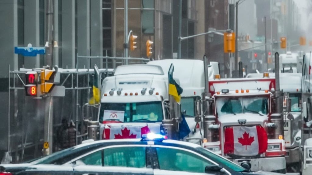 Snow-covered semi trucks with Canadian flags and a police car with flashing lights lined up on a city street.