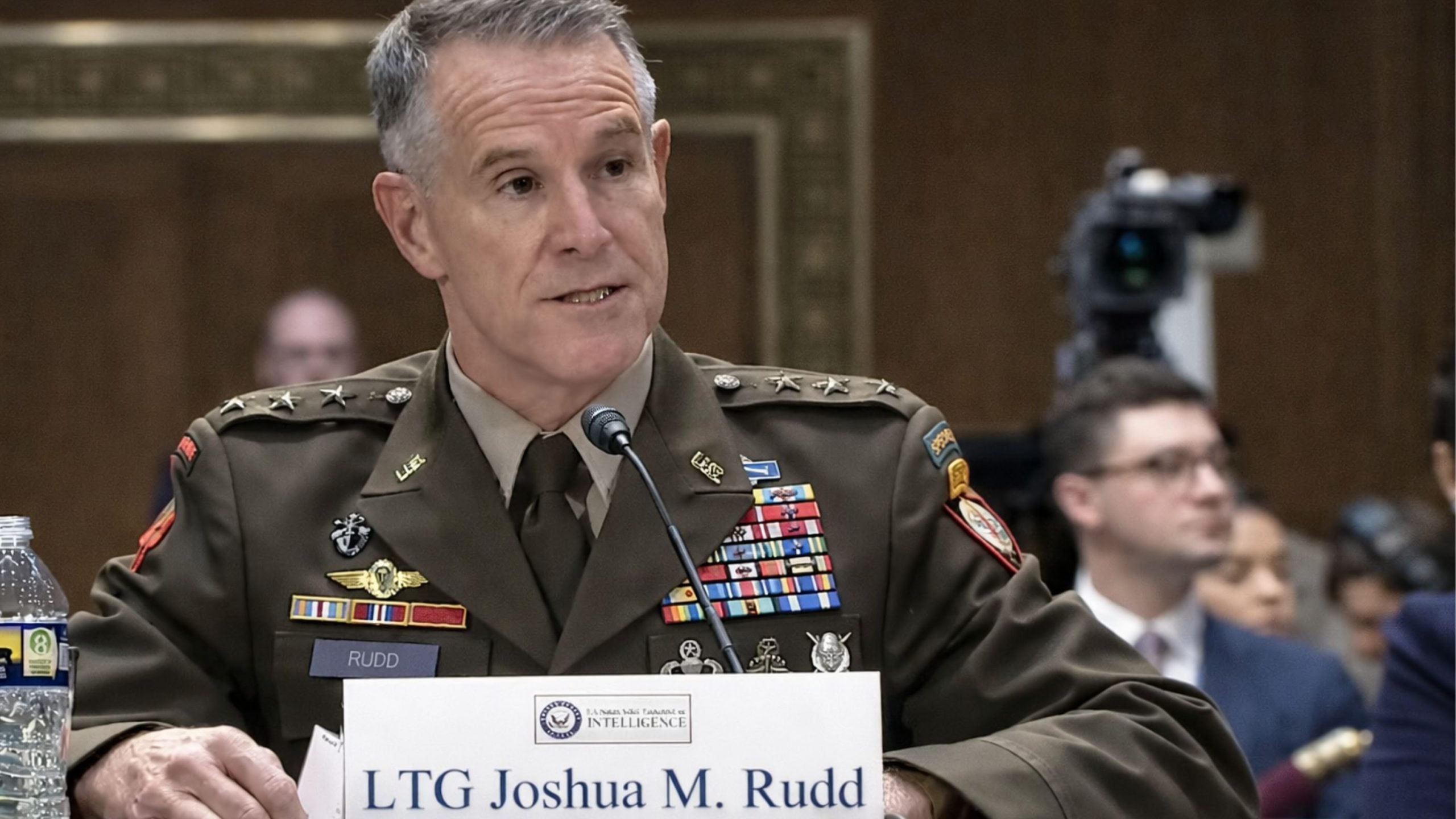 Uniformed senior U.S. Army officer with ribbons and medals speaking into a microphone at a hearing, name placard visible.