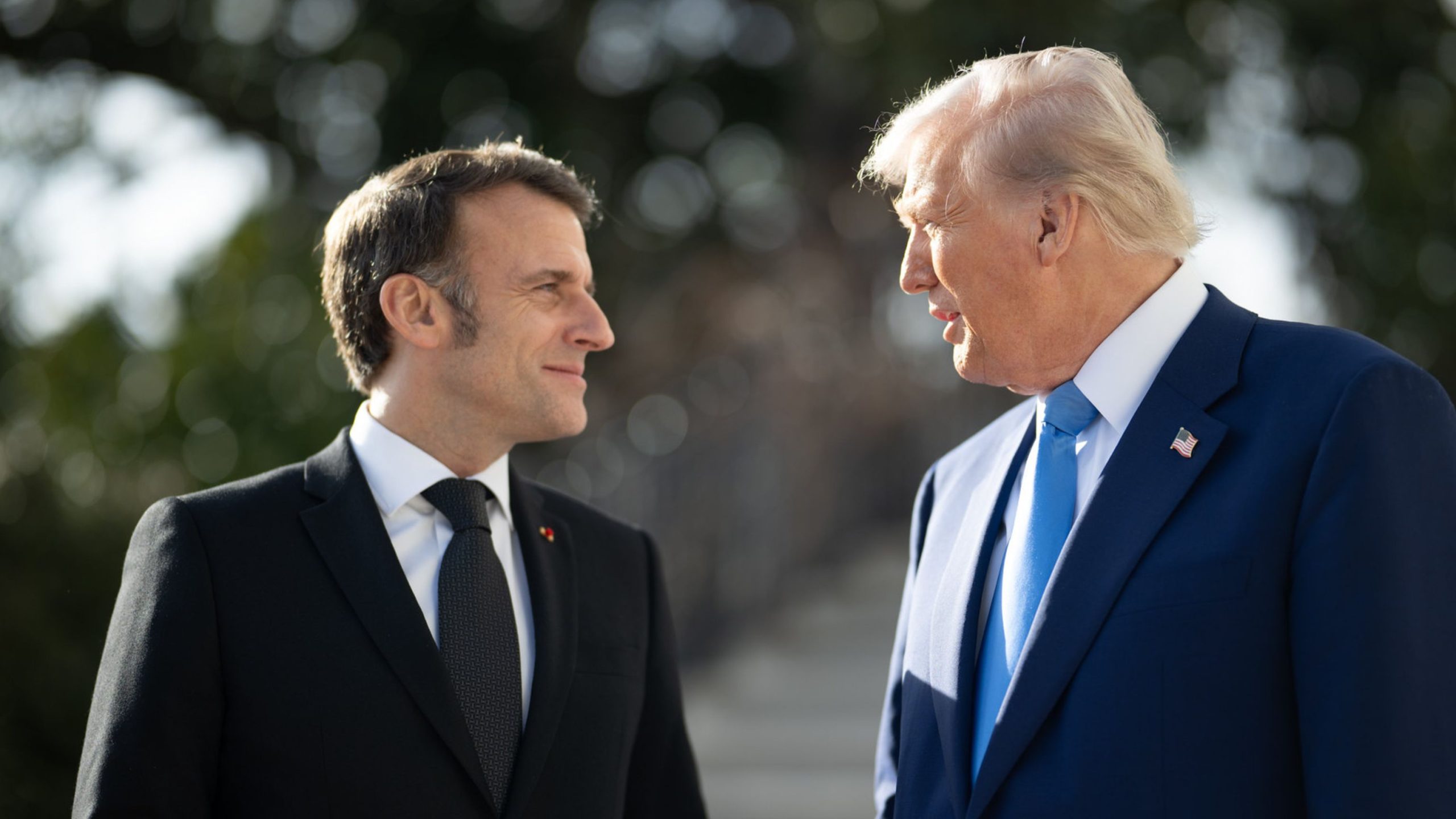 Macron and Trump in suits facing each other outdoors, one in black with a dark tie, the other in navy with a blue tie and a U.S. flag lapel pin.