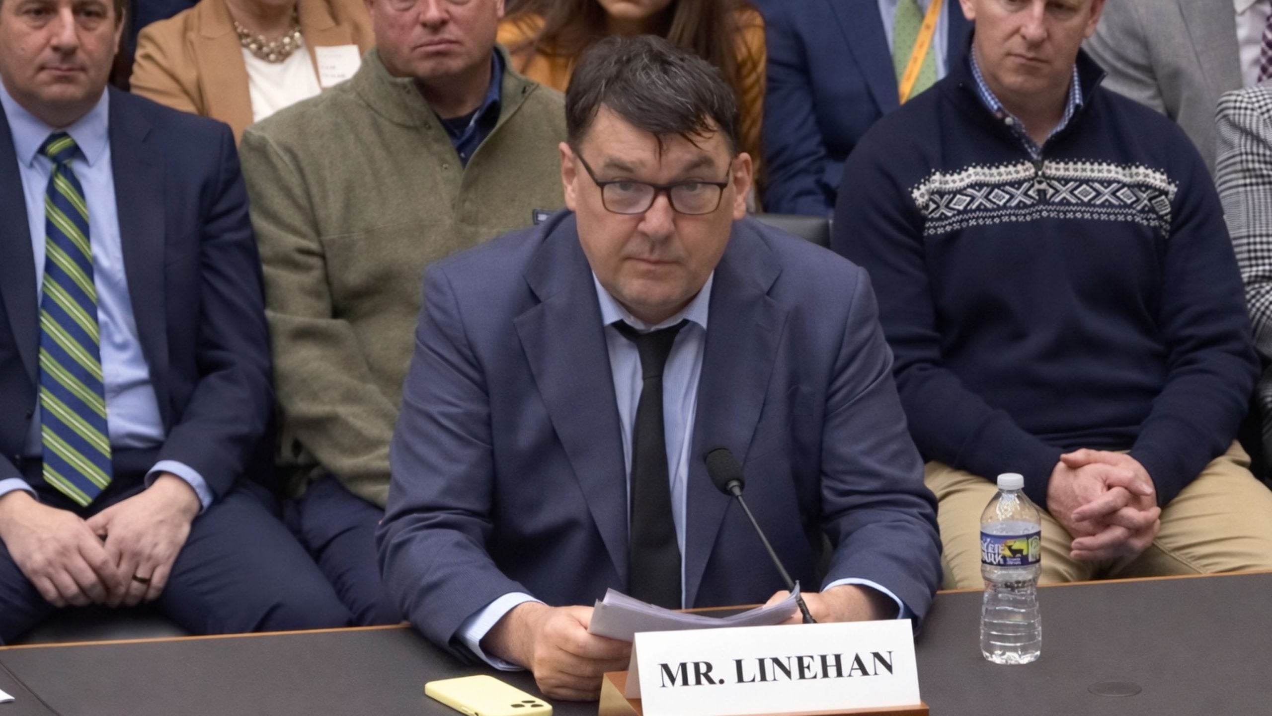 Linehan seated at a hearing table, holding papers in front of a nameplate and microphone.